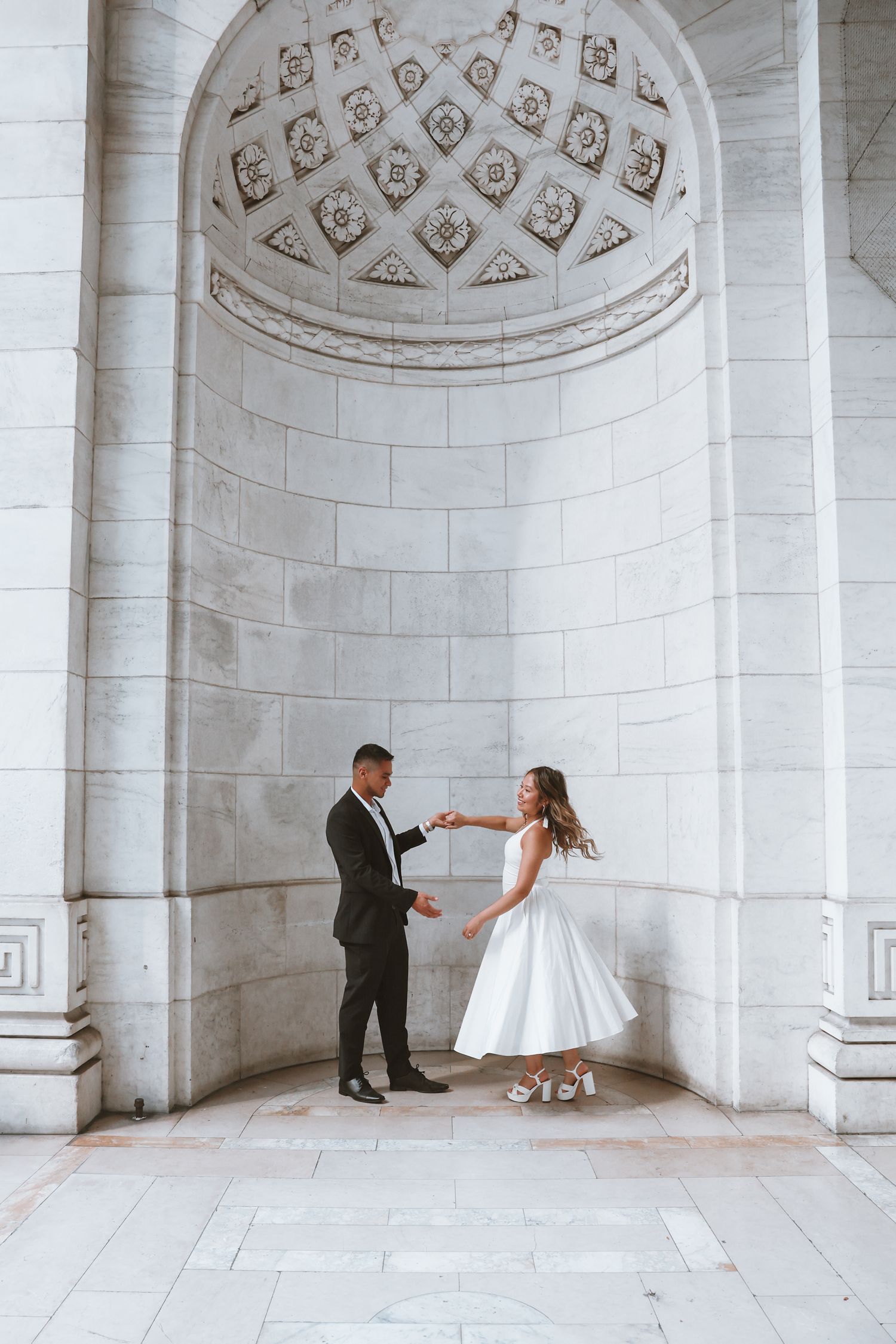 Bride in a white tea-length dress and groom in a dark suit dance hand in hand beneath a coffered marble arch for an elopement photo.