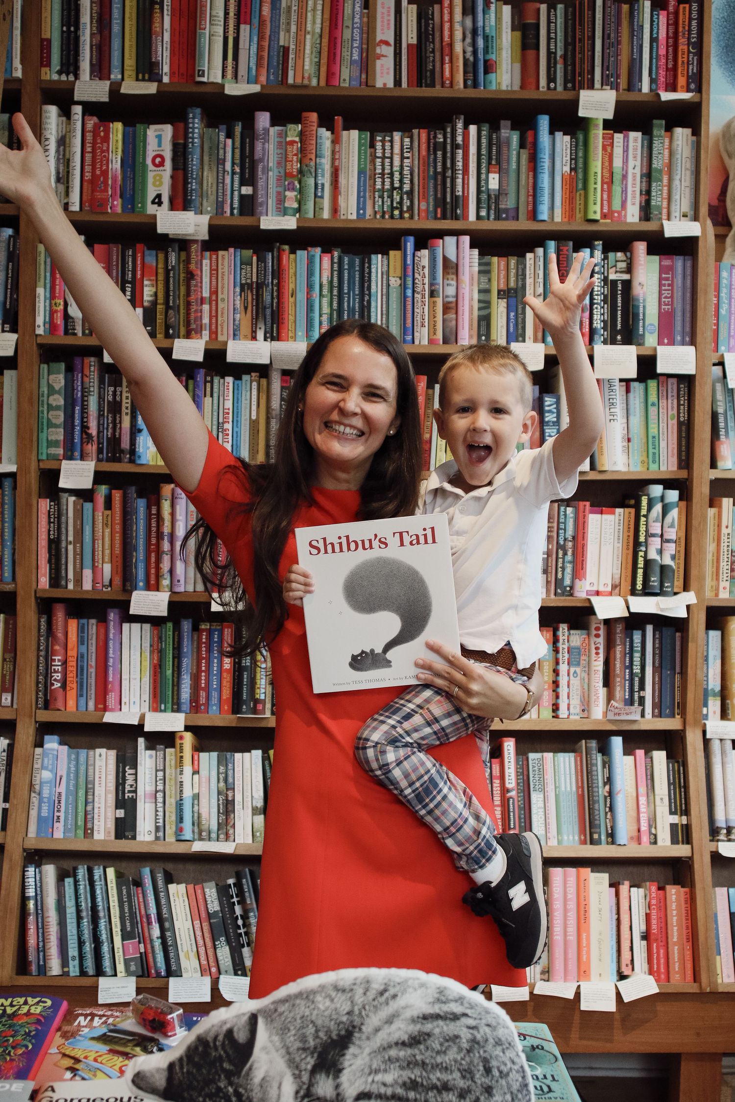 Smiling woman in red dress holding Shibu's Tail book with cheerful toddler in bookstore.