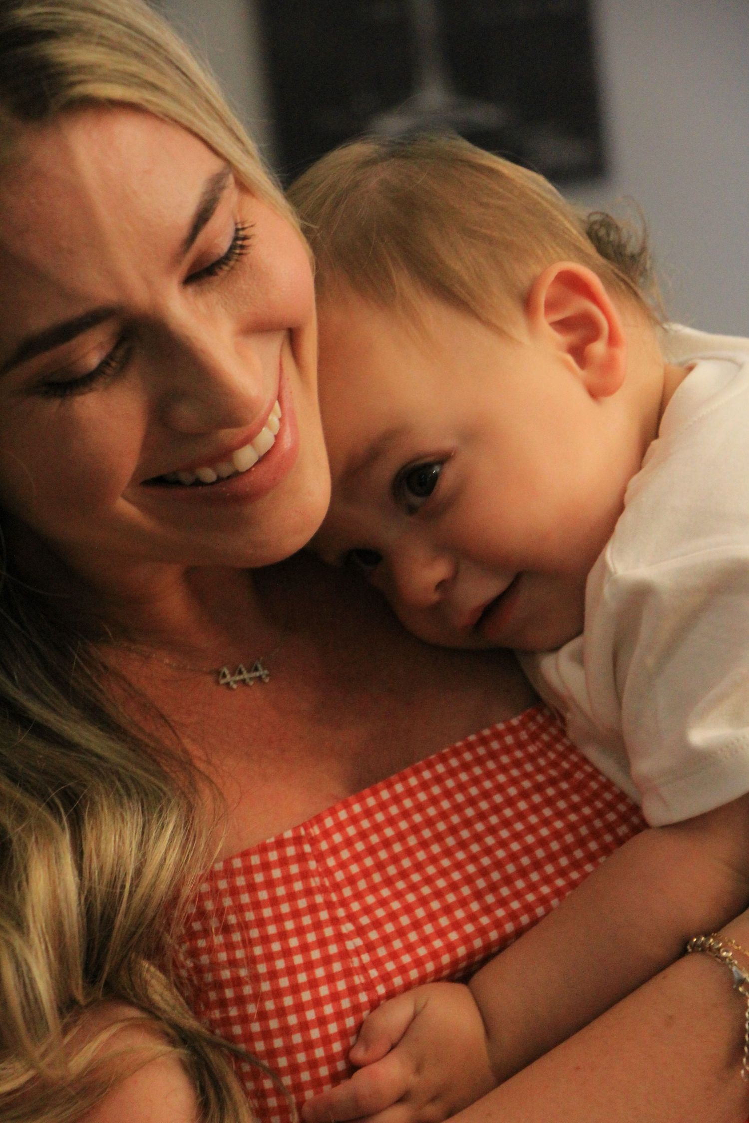 Smiling blonde woman in a red gingham dress holding a baby at an indoor celebration.