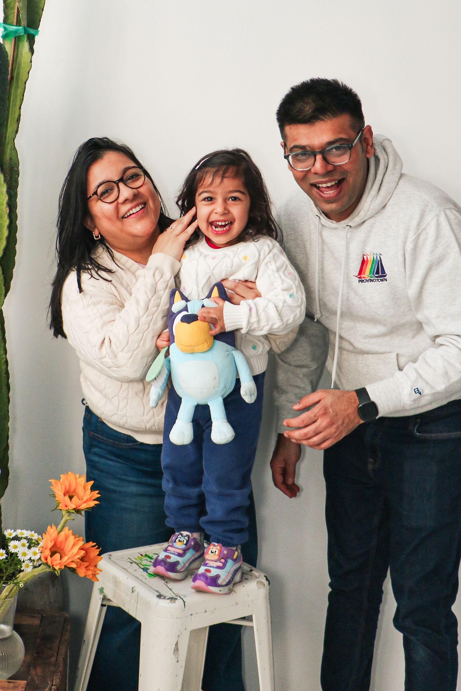Joyful family portrait with parents and toddler holding a Bluey stuffed toy standing on a white stool.