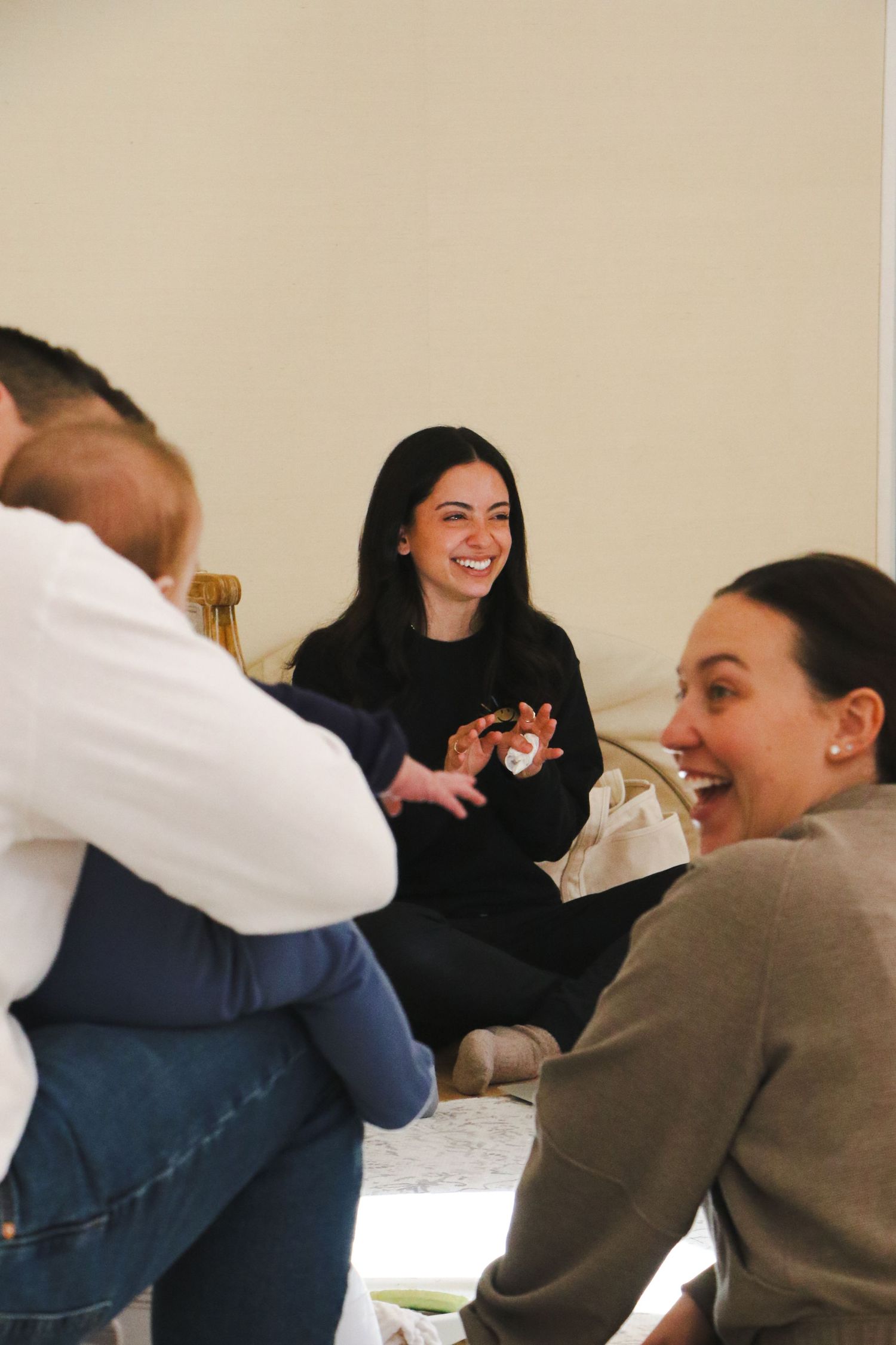 Young woman in black smiling warmly while sitting with a group of people holding a baby indoors.