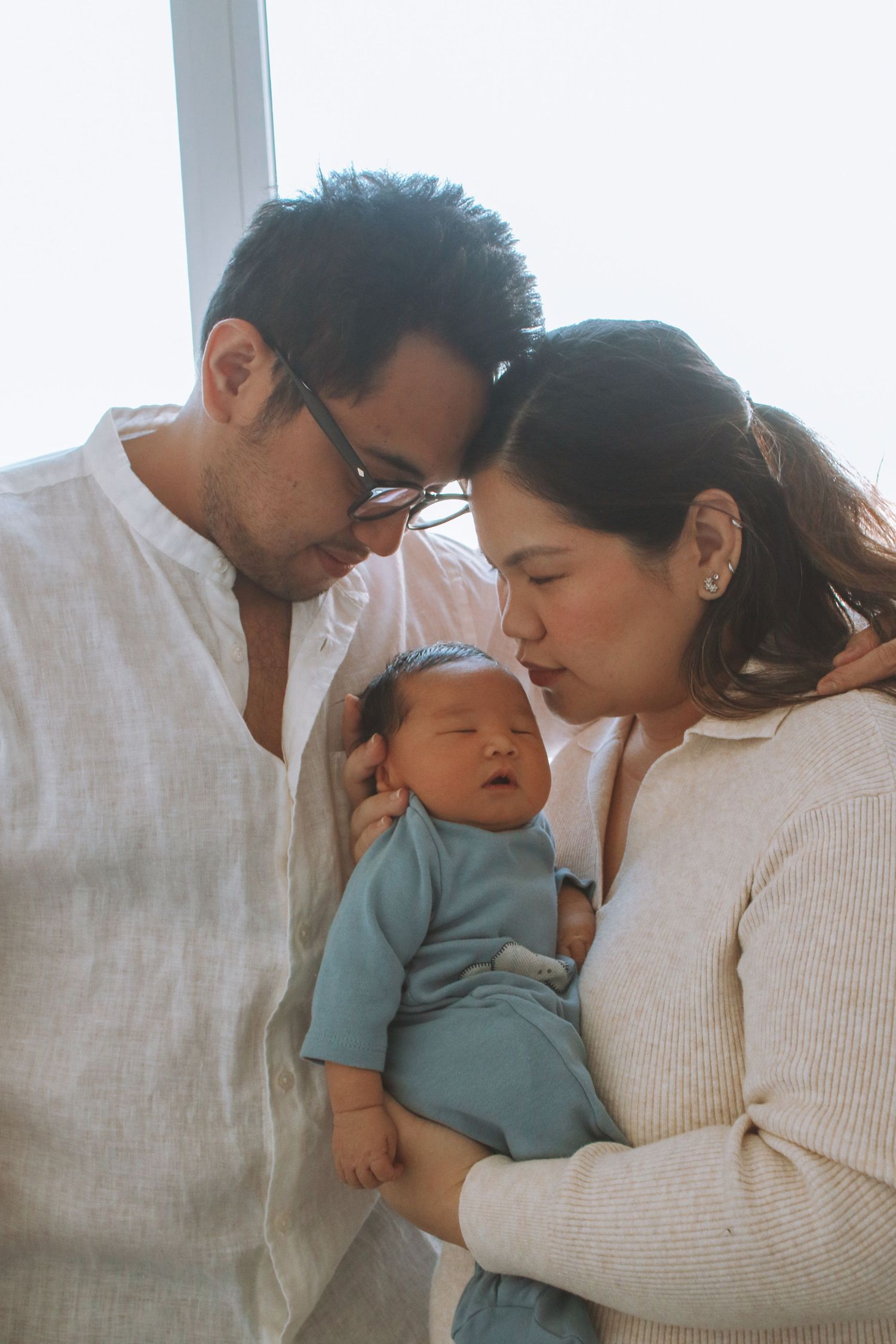 Loving parents lean in to kiss and cuddle their newborn baby in blue outfit.