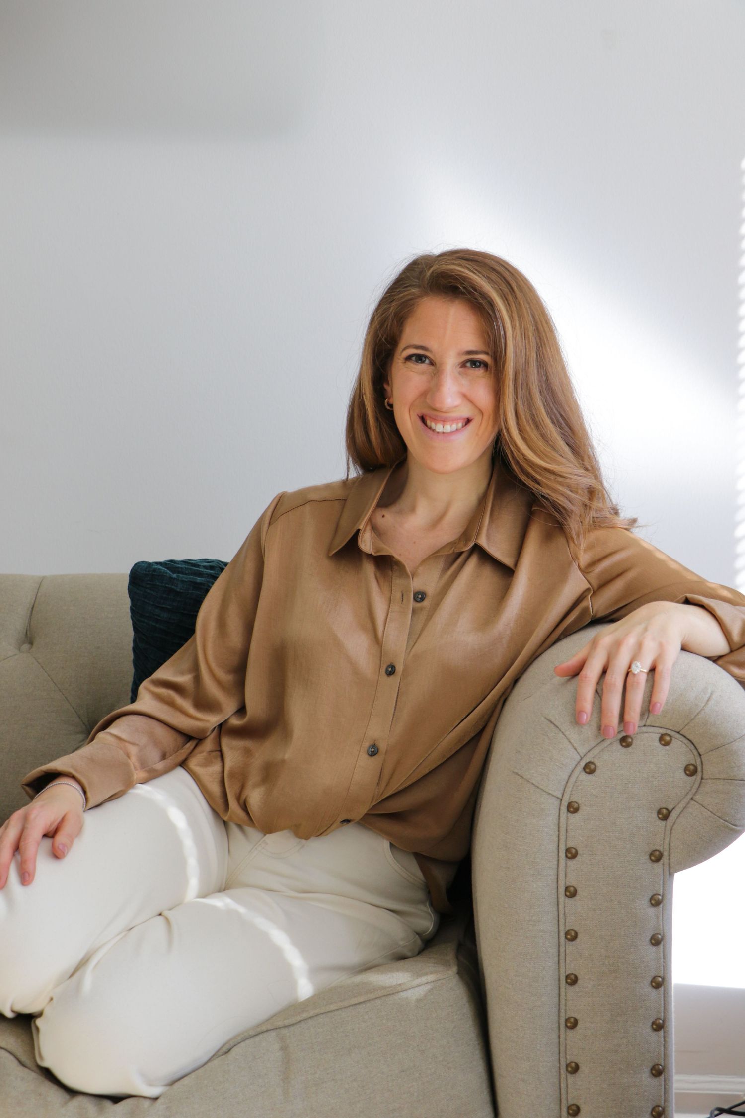 Smiling woman in tan blouse and white pants relaxing on gray tufted sofa.