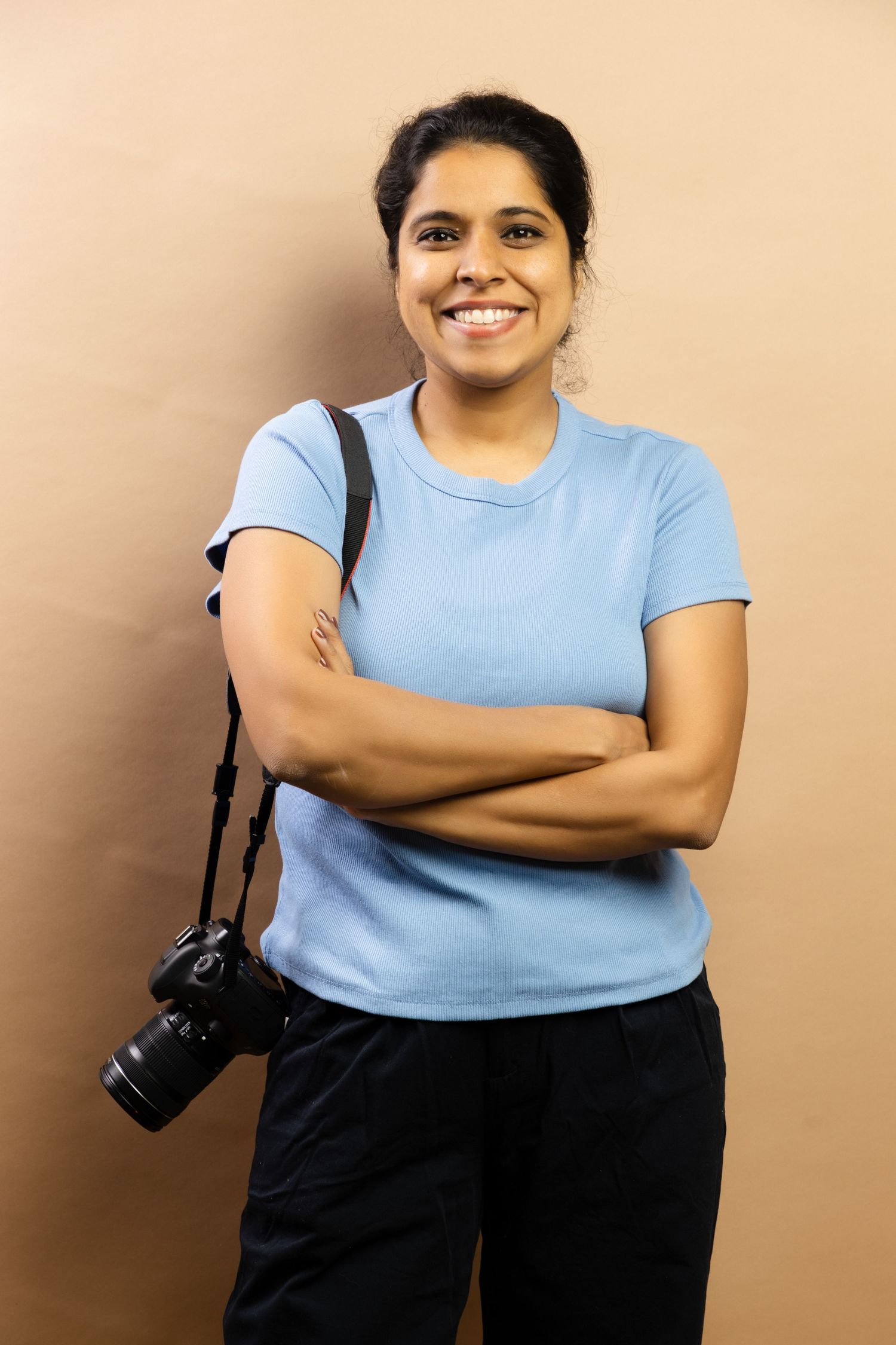 Confident Indian female photographer smiling with arms crossed, camera hanging by strap, beige background.
