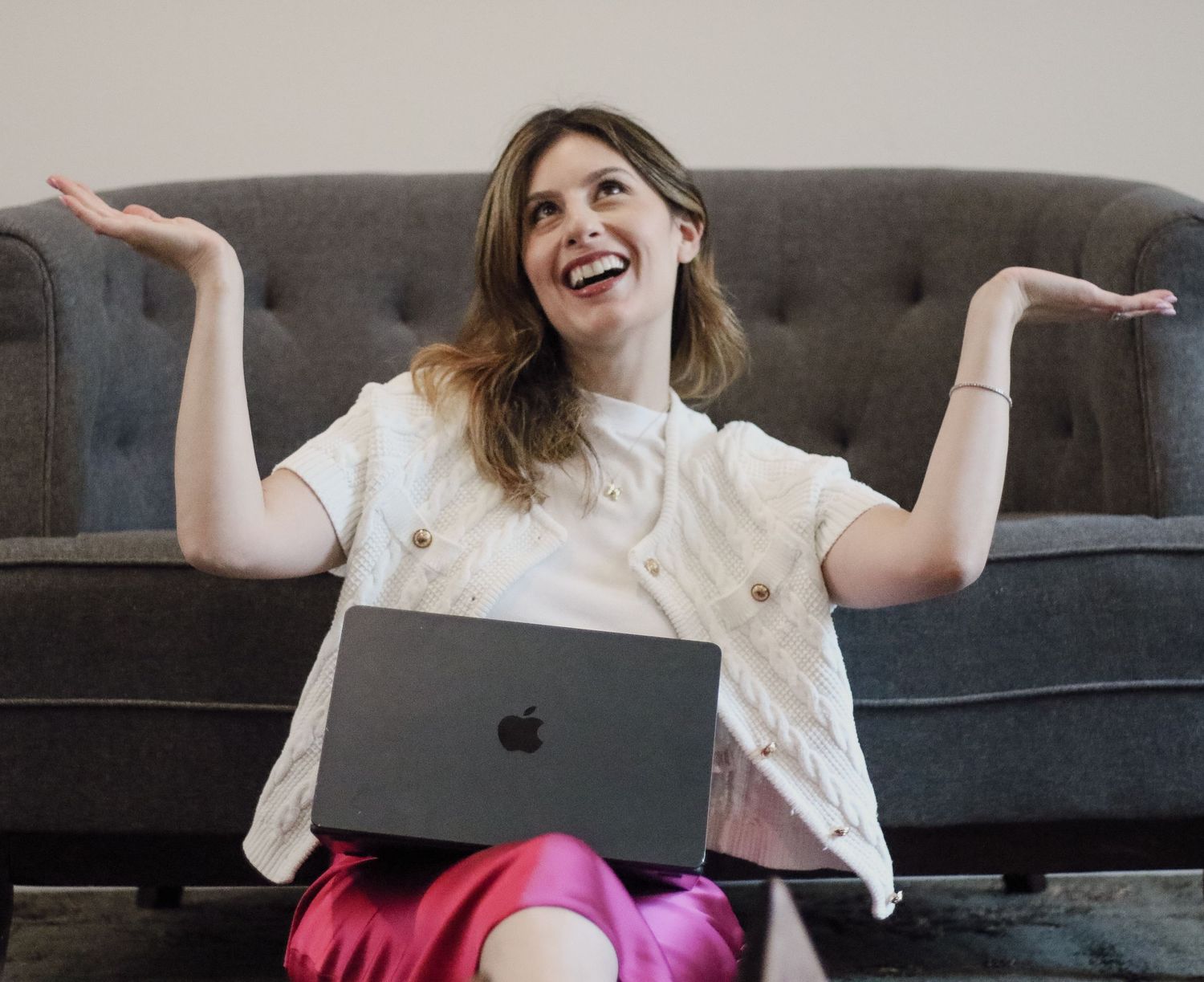 Woman laughing with hands raised sitting on floor with Apple MacBook laptop against gray sofa.