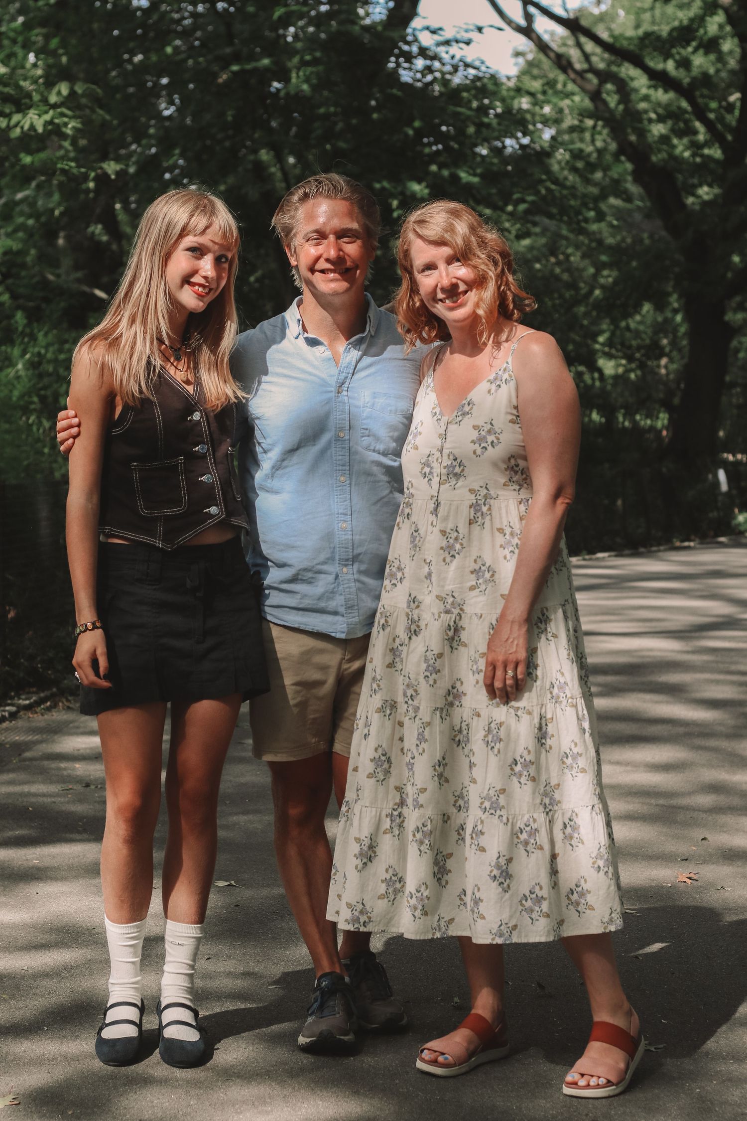 Family of three smiling together on sunny tree-lined path in park.