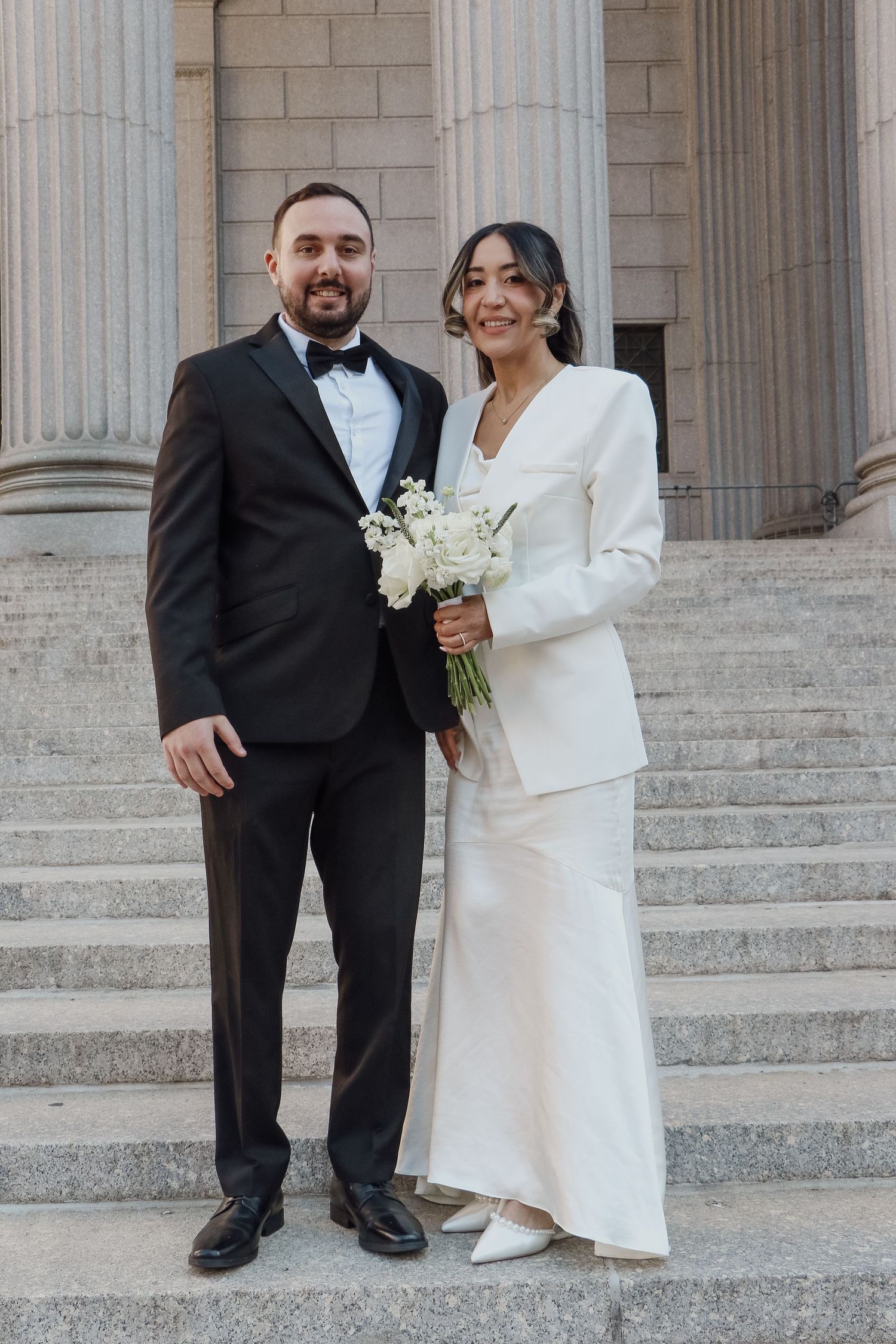 Bride in white blazer gown and groom in black tuxedo pose together on stone steps holding bouquet.