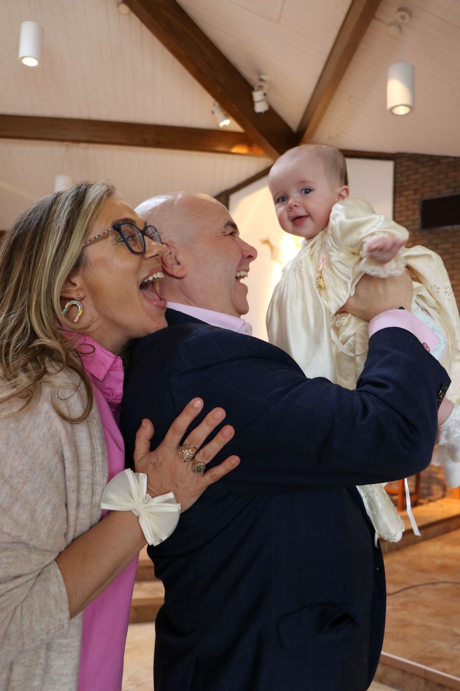 Joyful couple holding smiling baby in christening gown inside church.