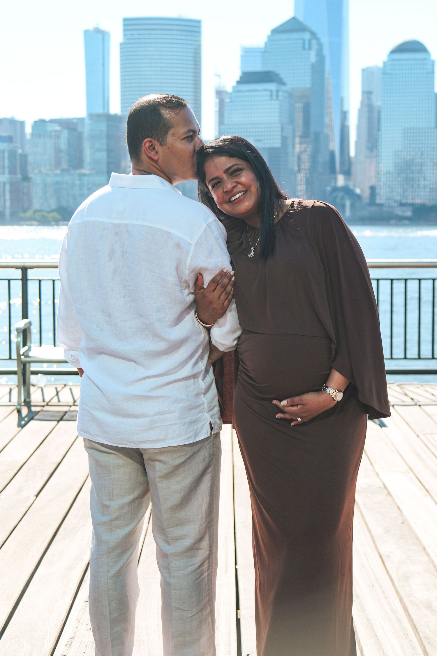 Pregnant couple posing on waterfront deck with NYC skyline backdrop, man kissing woman's cheek as she smiles.