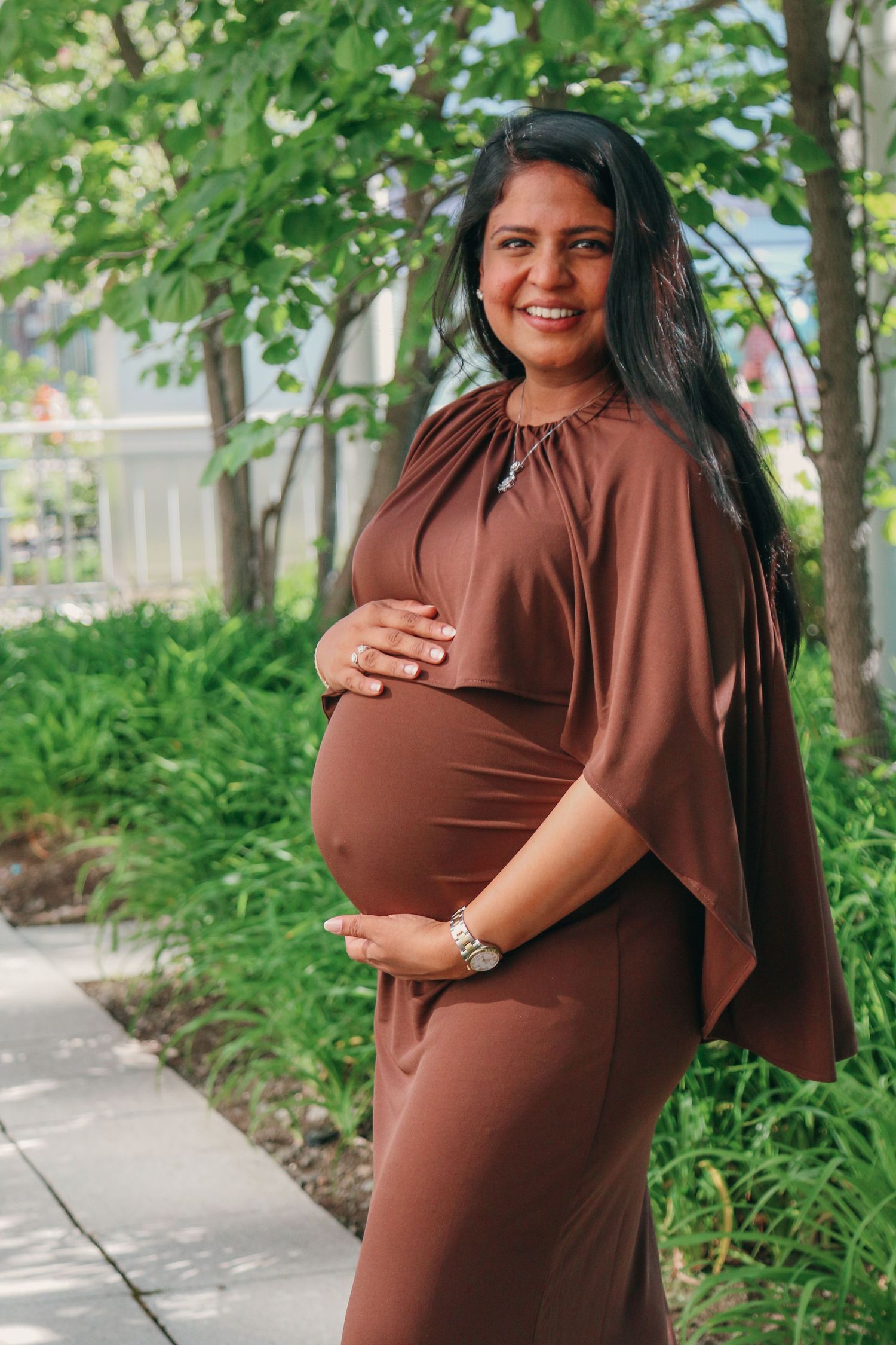 Smiling pregnant woman in brown dress cradling baby bump outdoors among green trees and lush garden path.