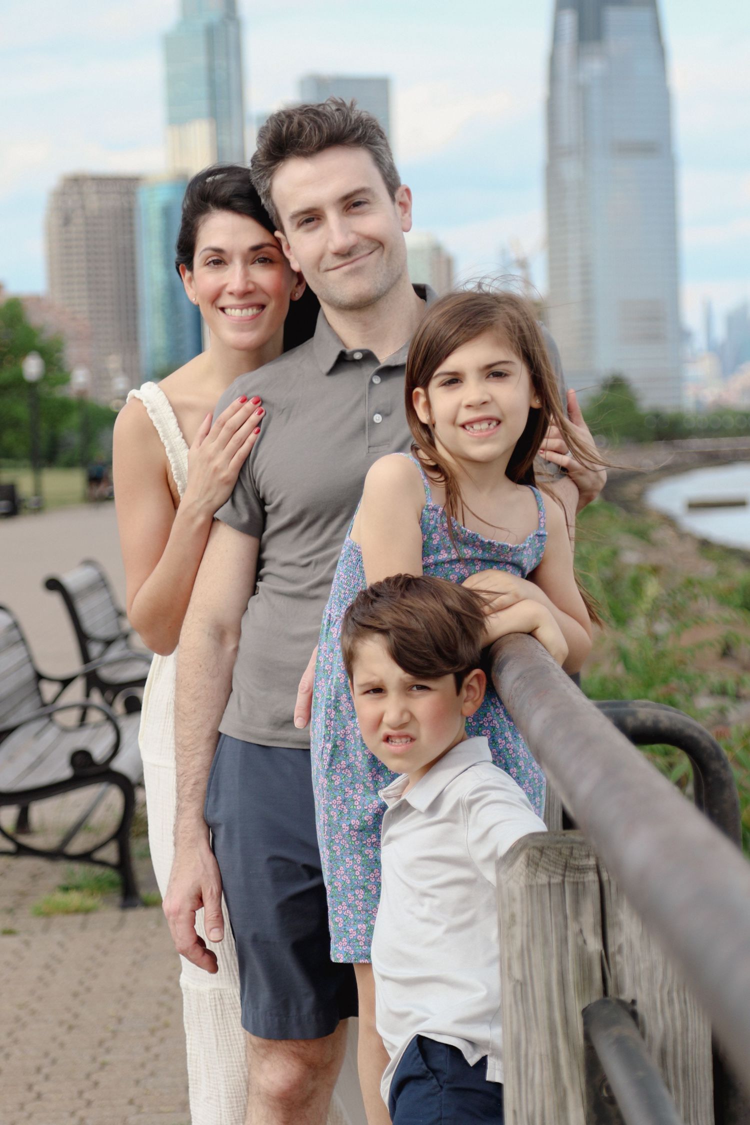 A happy family of four poses by a railing with a city skyline in the background on a cloudy day.