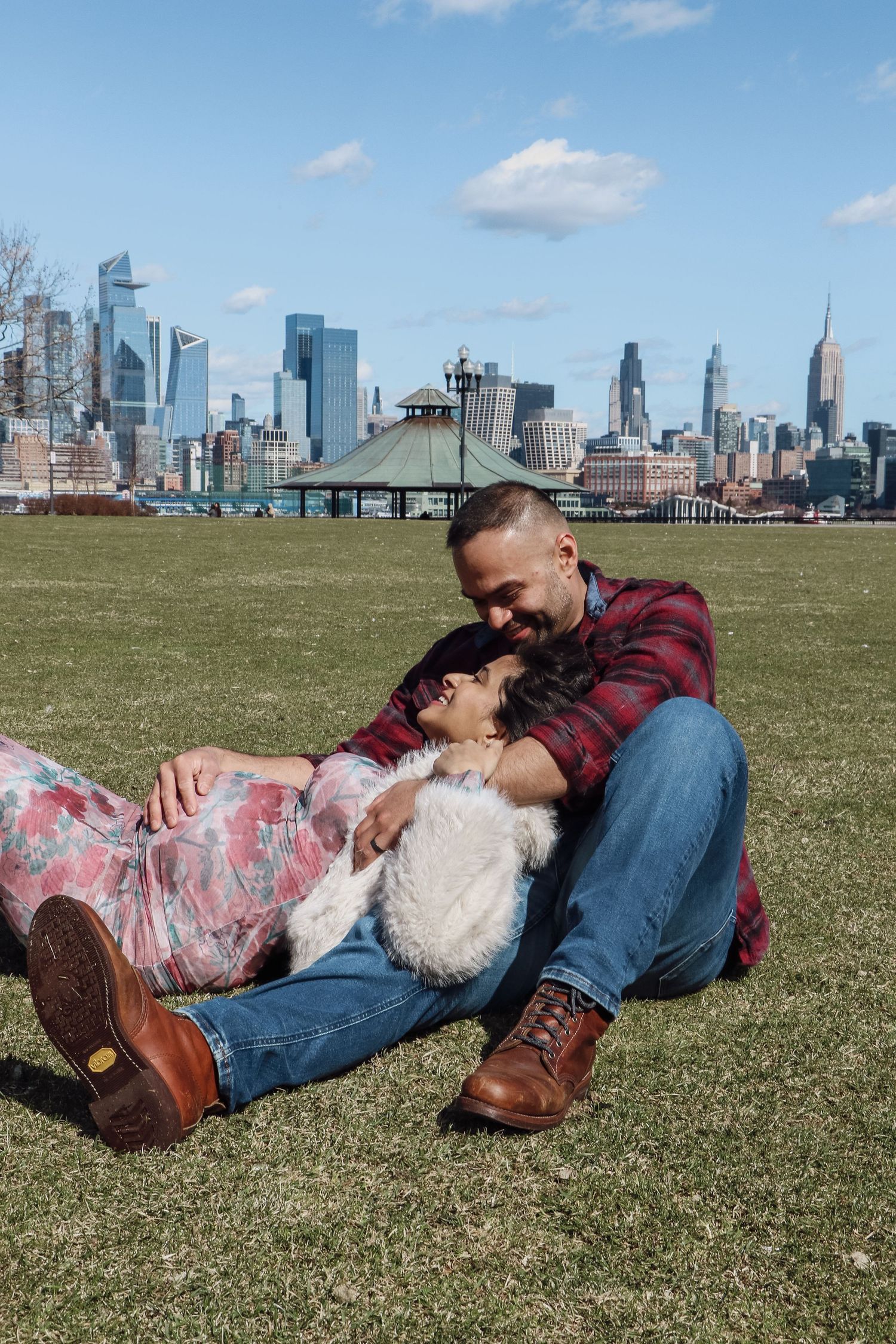 Couple relaxing on grass in park with NYC skyline backdrop, woman lying in man's lap smiling.