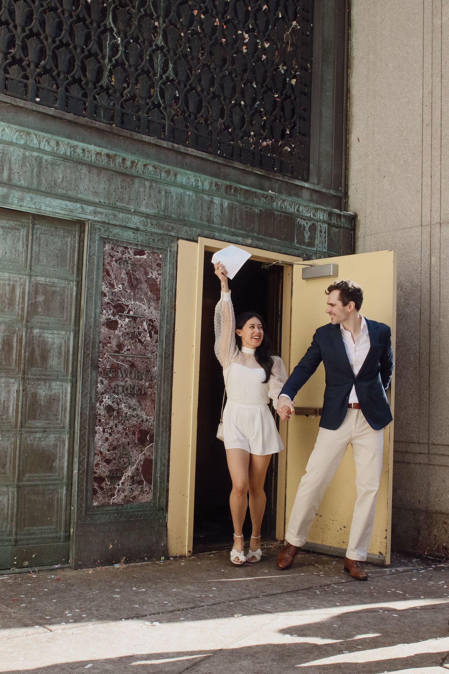 Joyful couple exits NYC City Clerk office, bride in white romper raising paper, groom in navy blazer holding hands.