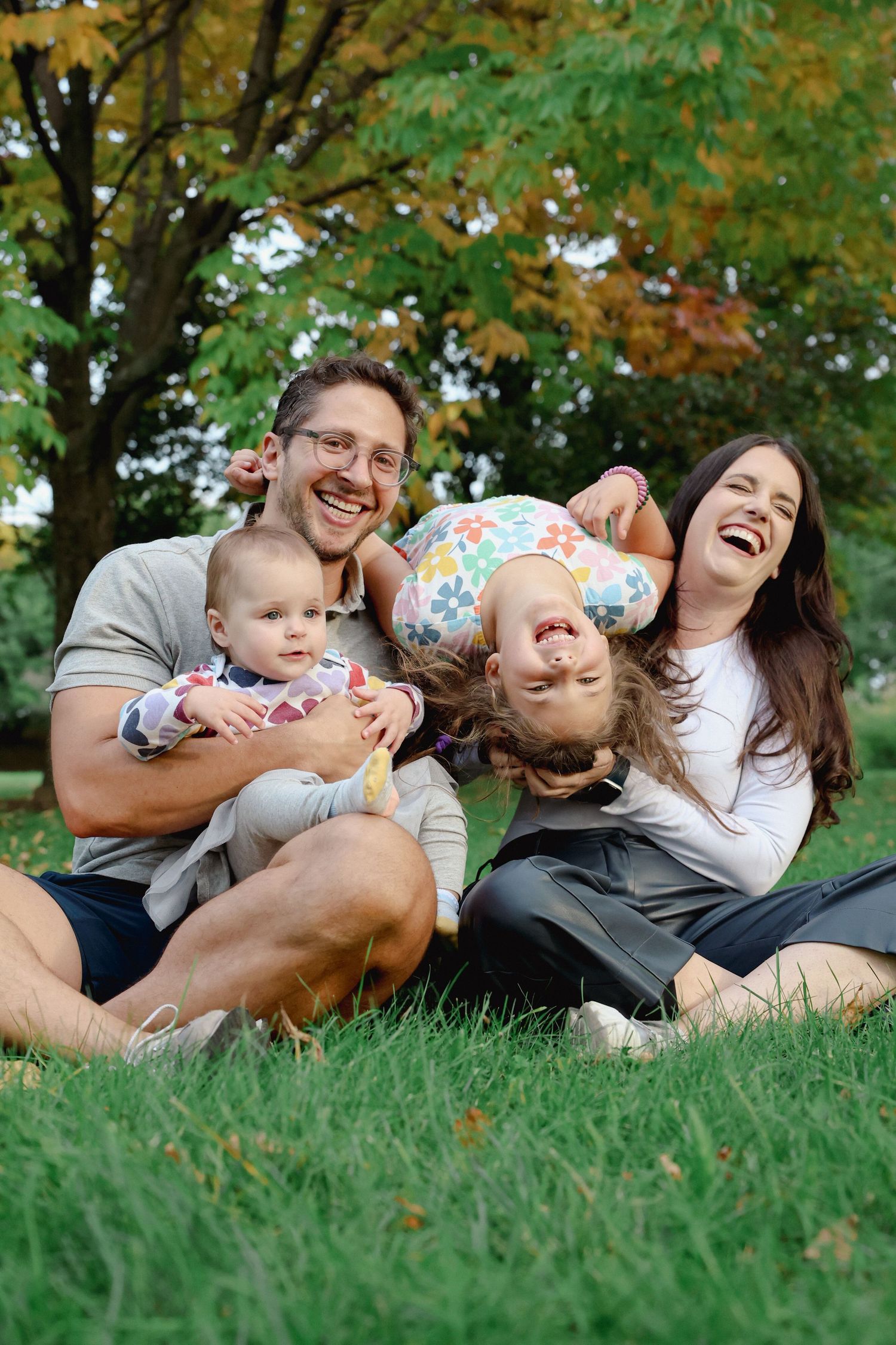A laughing family of four sits on a grassy lawn beneath autumn trees as parents hold their baby and a giggling daughter upside down.