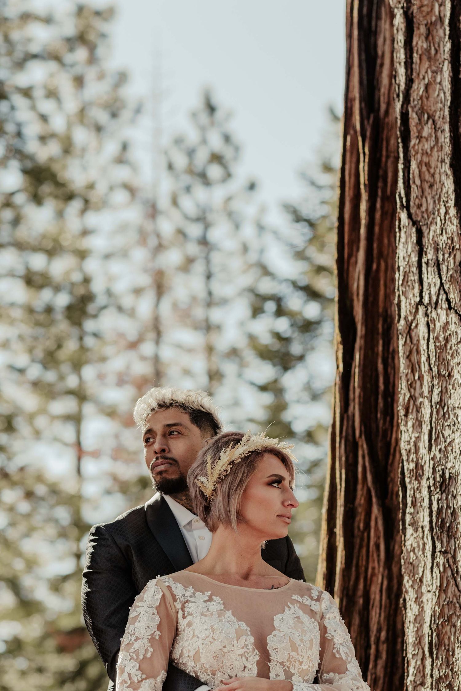 groom stand behind bride embracing her. they look in opposite directions. pine trees in the background