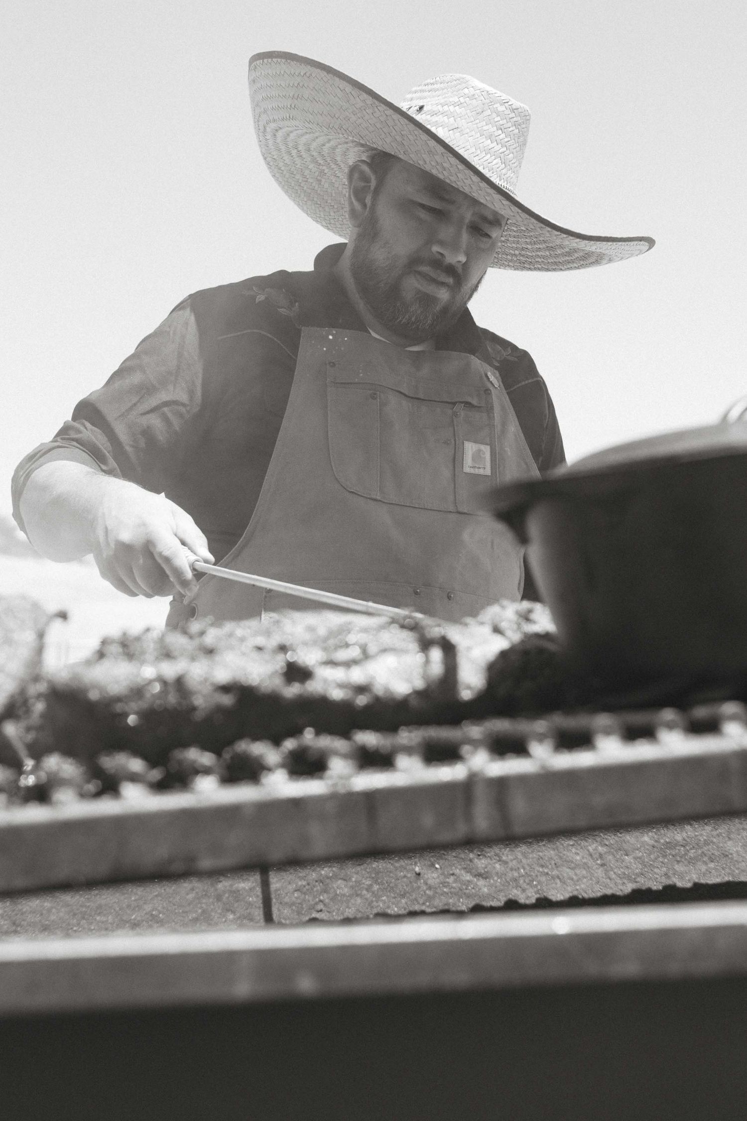 b+w photo of chef wearing large sombrero while checking meat on the grill