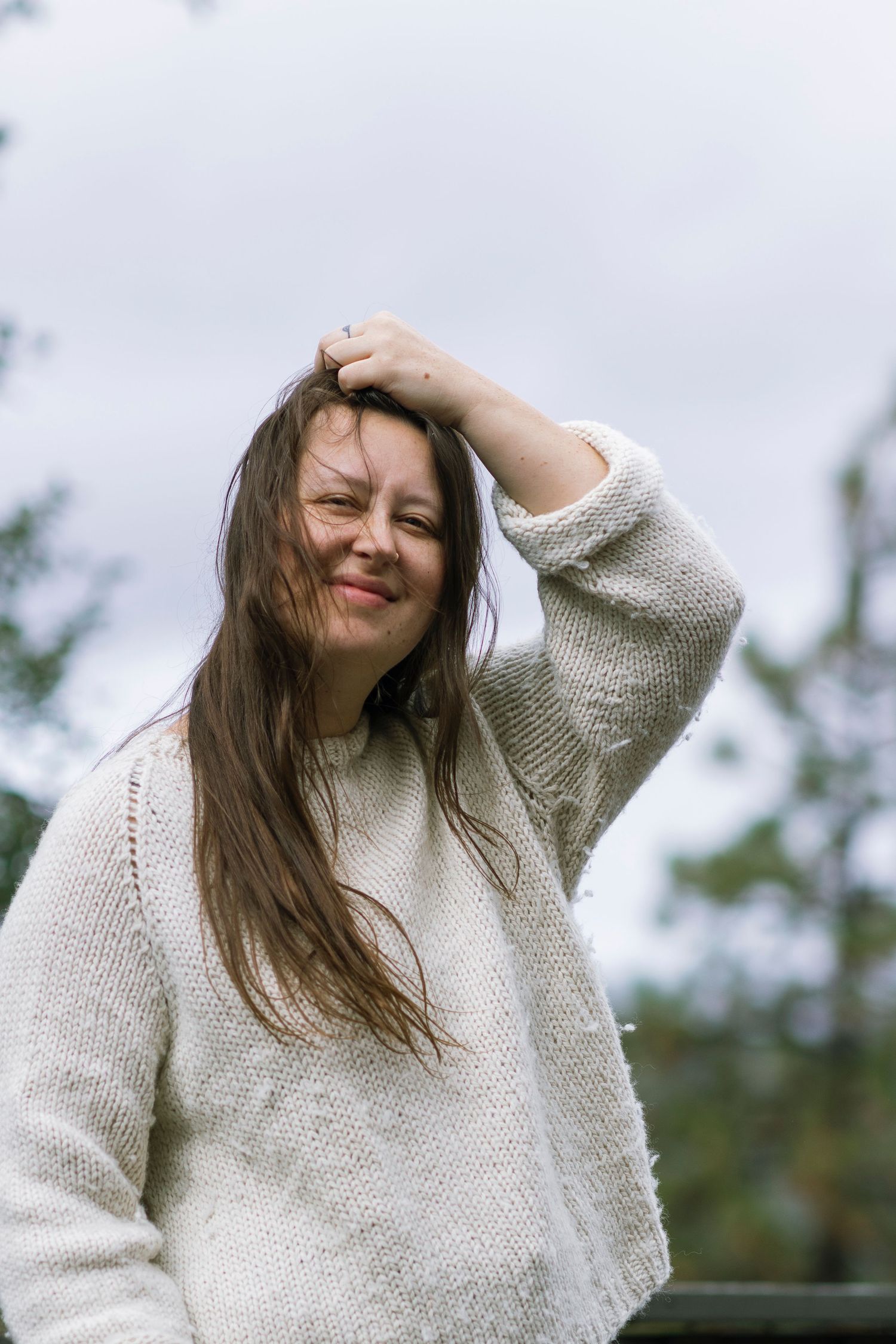 woman wearing white knit sweater stands and pushes hair out of face with hand.
