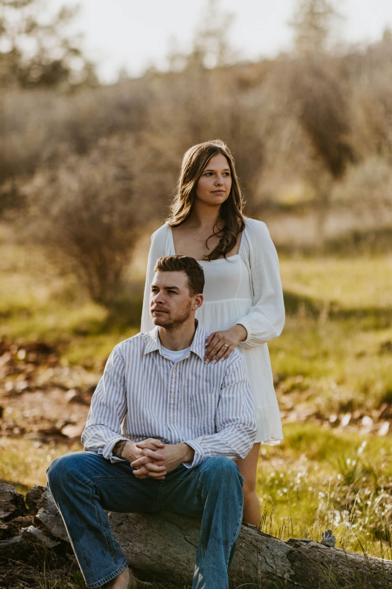 man sits on log, woman stands behind him with one hand resting on his shoulder. they look in different directions