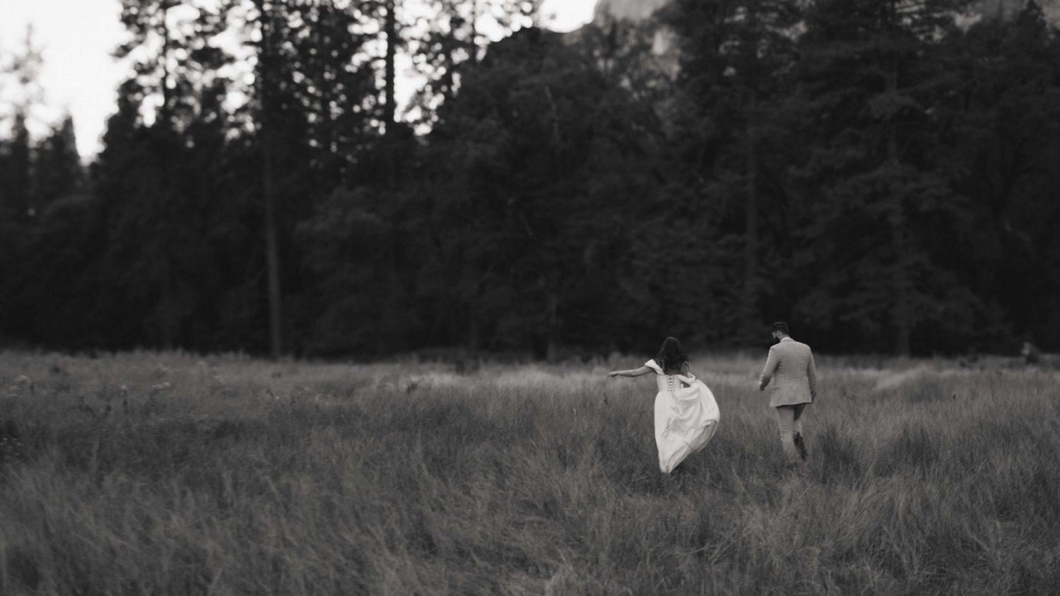 b+w photo of bride and groom running away in a grassy field in yosemite