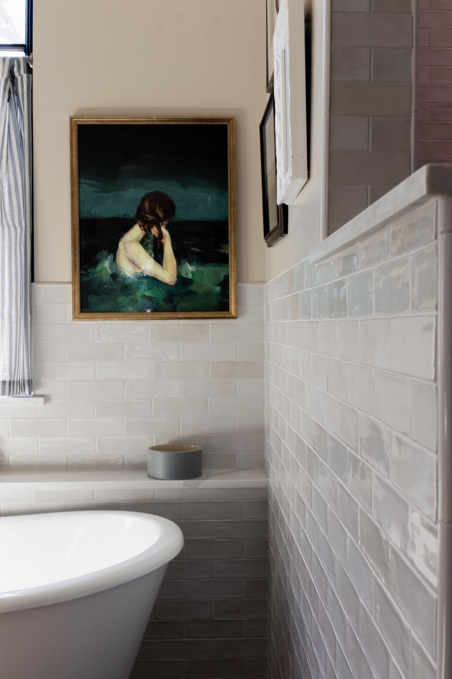 ceramic bathtub and white tiled wall in corner of bathroom. blue and green artwork hangs on wall.