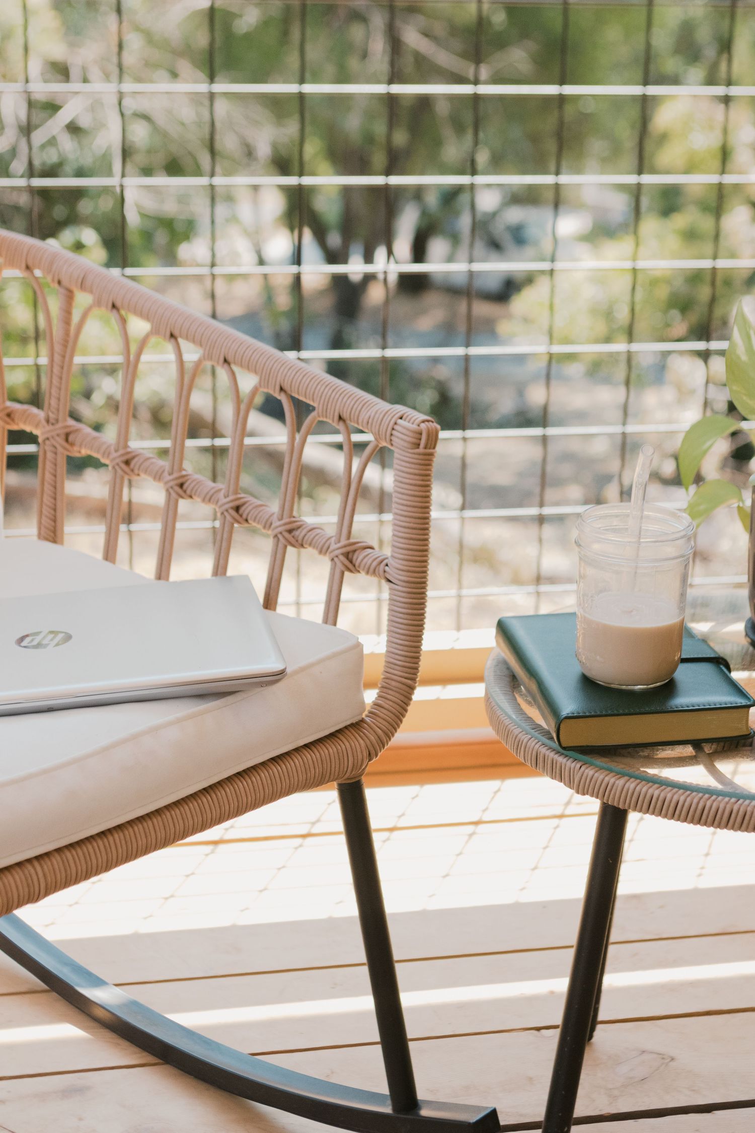 laptop sits on a rattan rocking chair. book sits on side table with glass of iced coffee on top