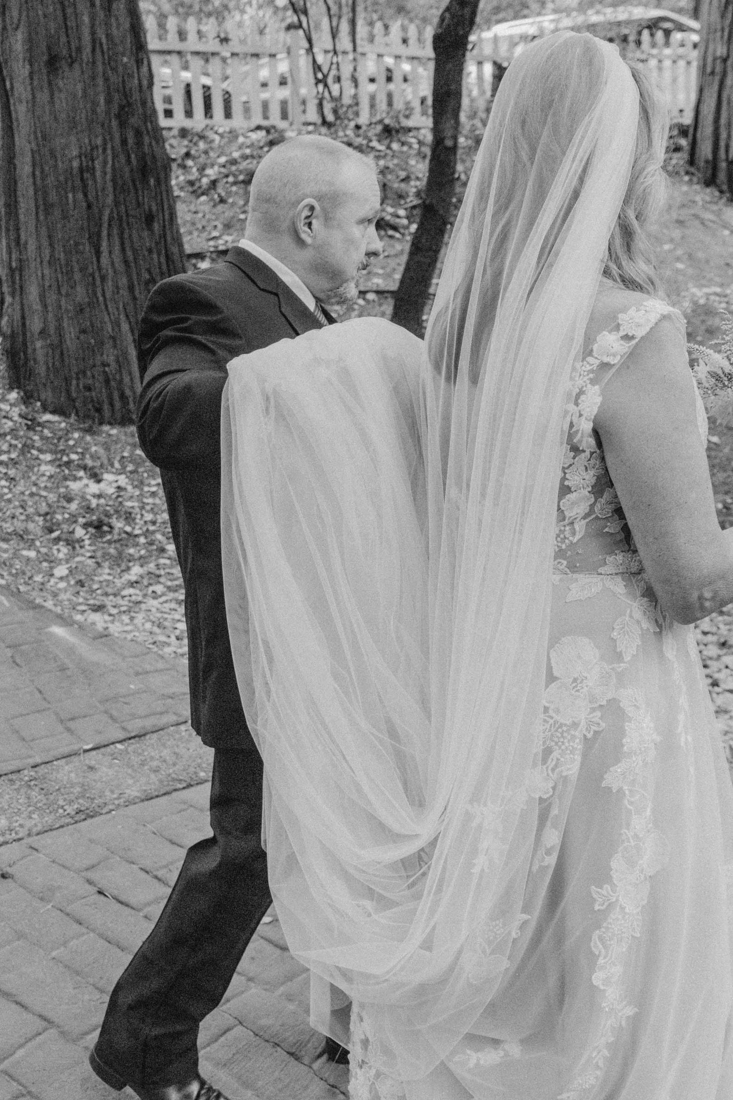 black and white image of bride and groom walking to reception. groom carries long train of bride's dress