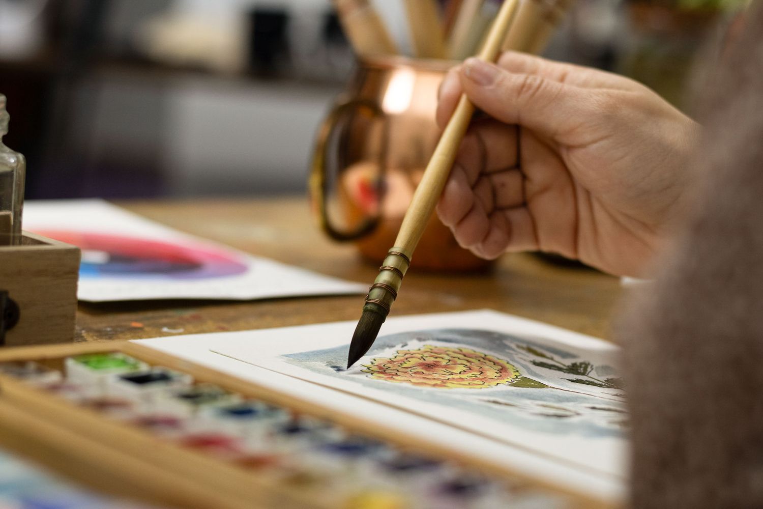 artist hand holding brush painting vibrant watercolor floral art on table with paints palette and brushes natural light creative portrait