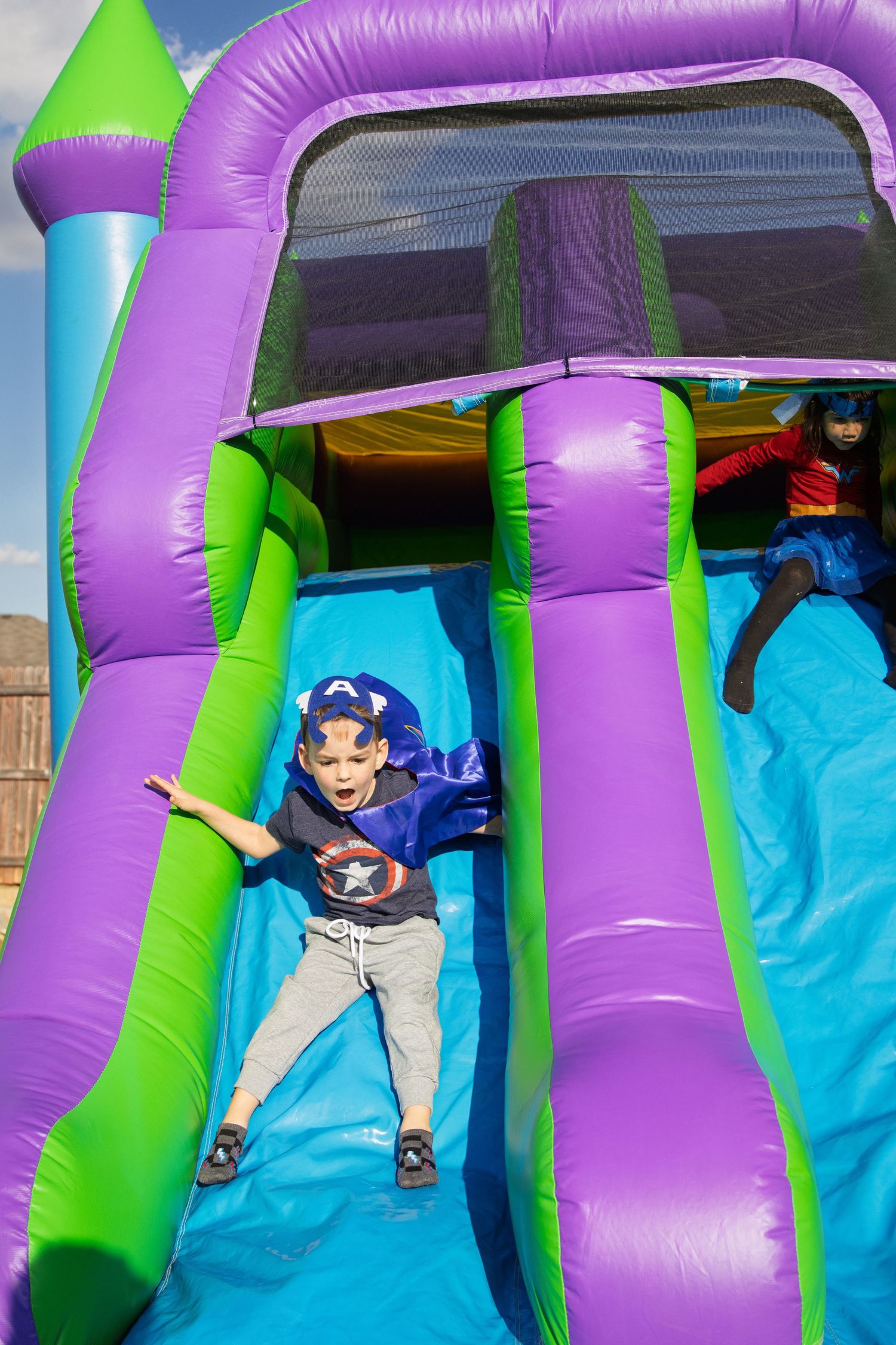 young boy in Captain America shirt sliding down inflatable bounce house slide outdoors at birthday party portrait photography natural light