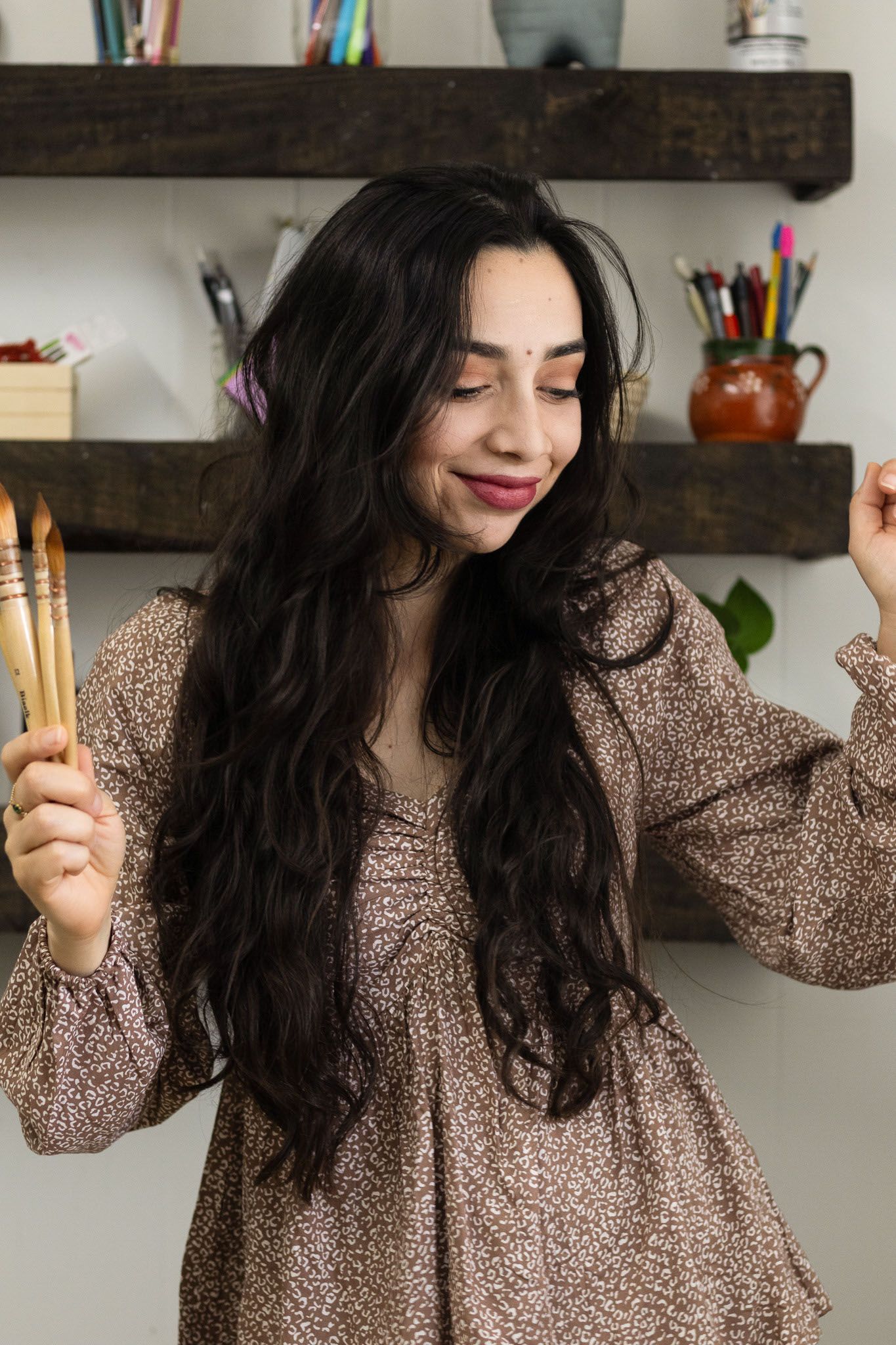 female artist holding paintbrushes smiling in creative studio portrait photo with shelves art supplies and studio light