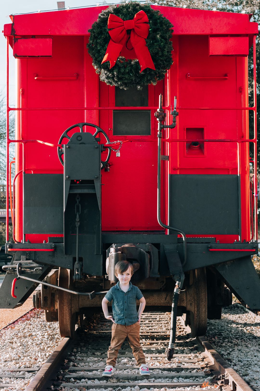 Young boy standing on railroad track in front of a vintage red caboose decorated with a Christmas wreath during a family photography session