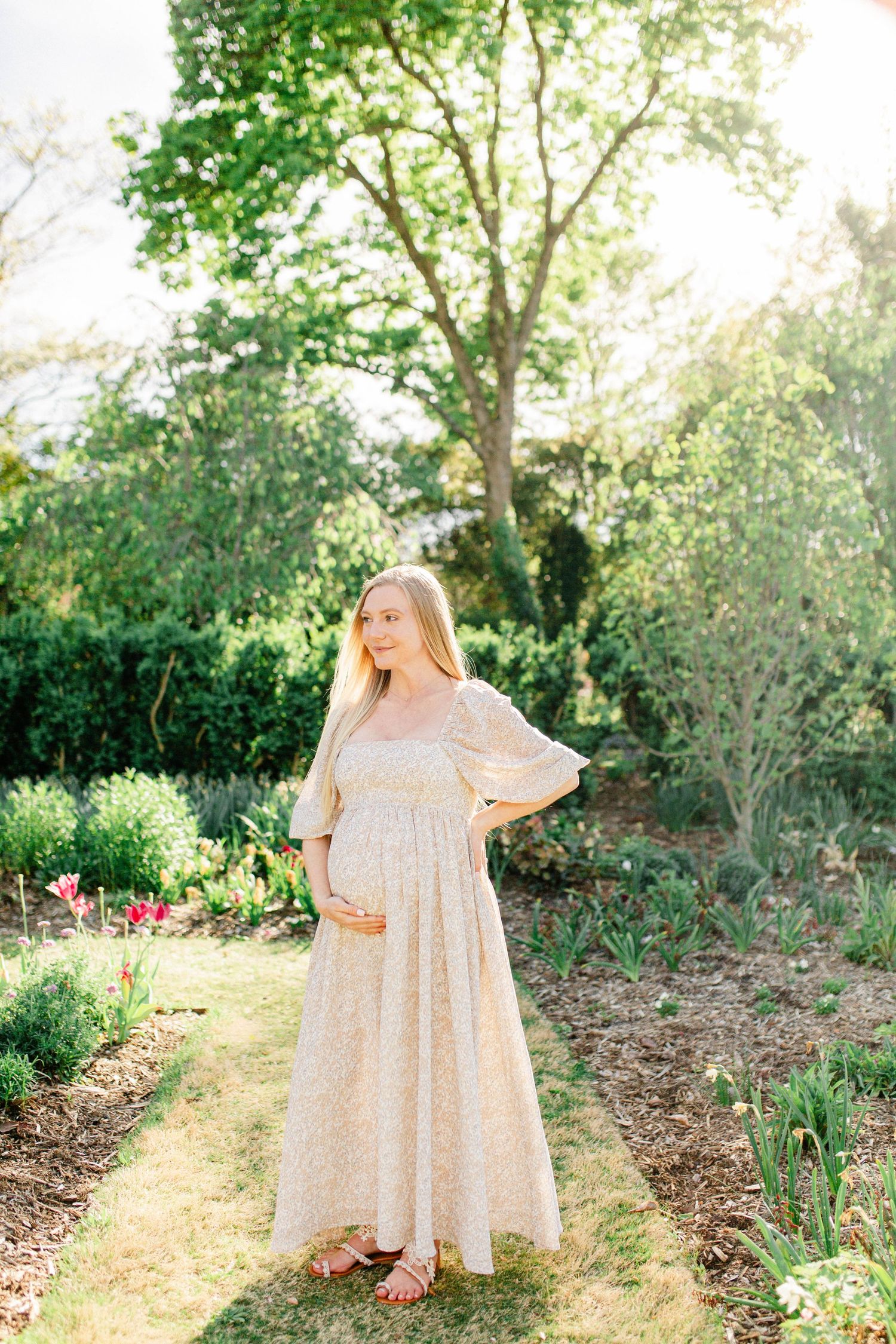 Woman in flowing cream dress poses in sunlit garden setting.