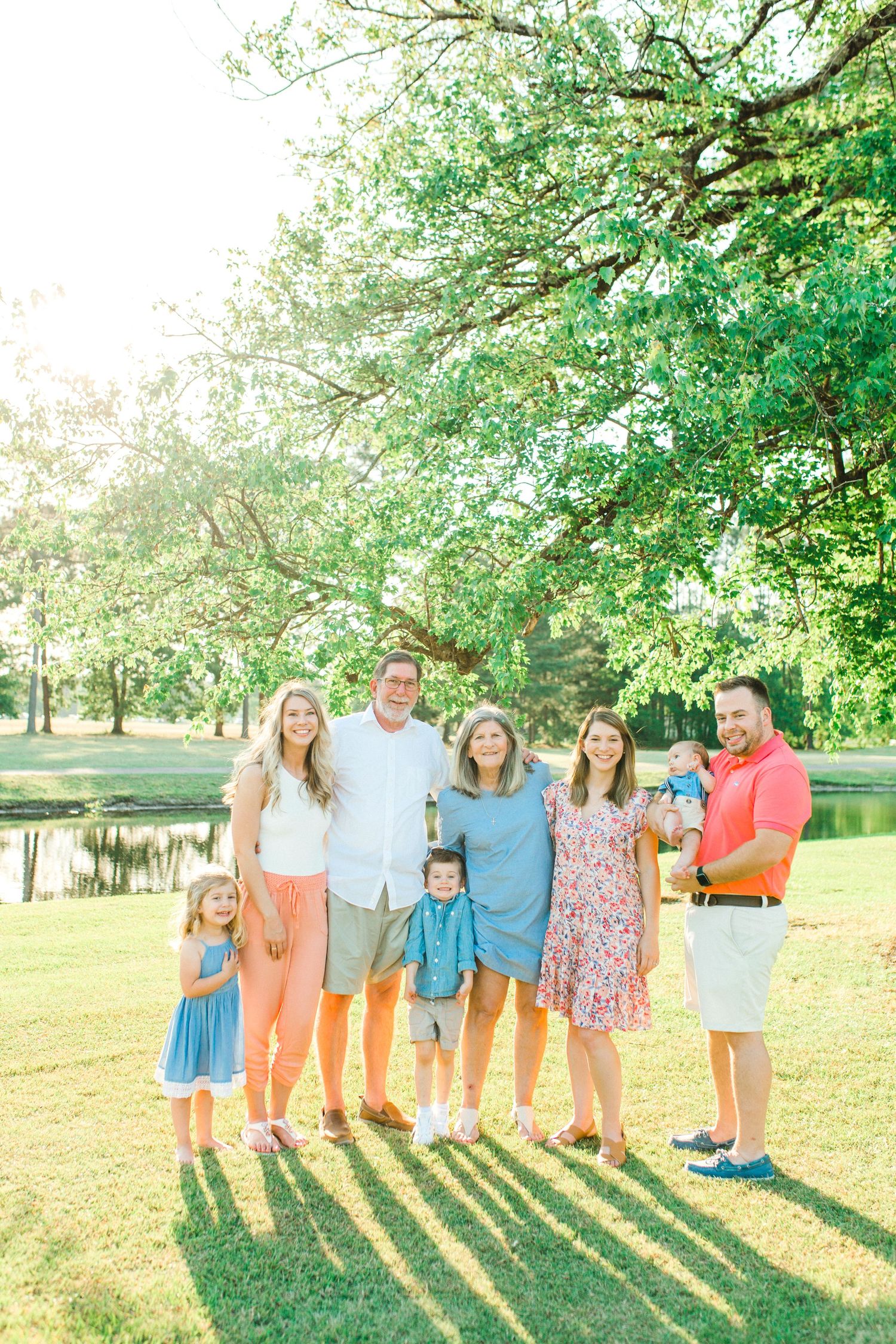 Family group poses together on grassy lawn during golden hour with long shadows cast behind them in summer setting.