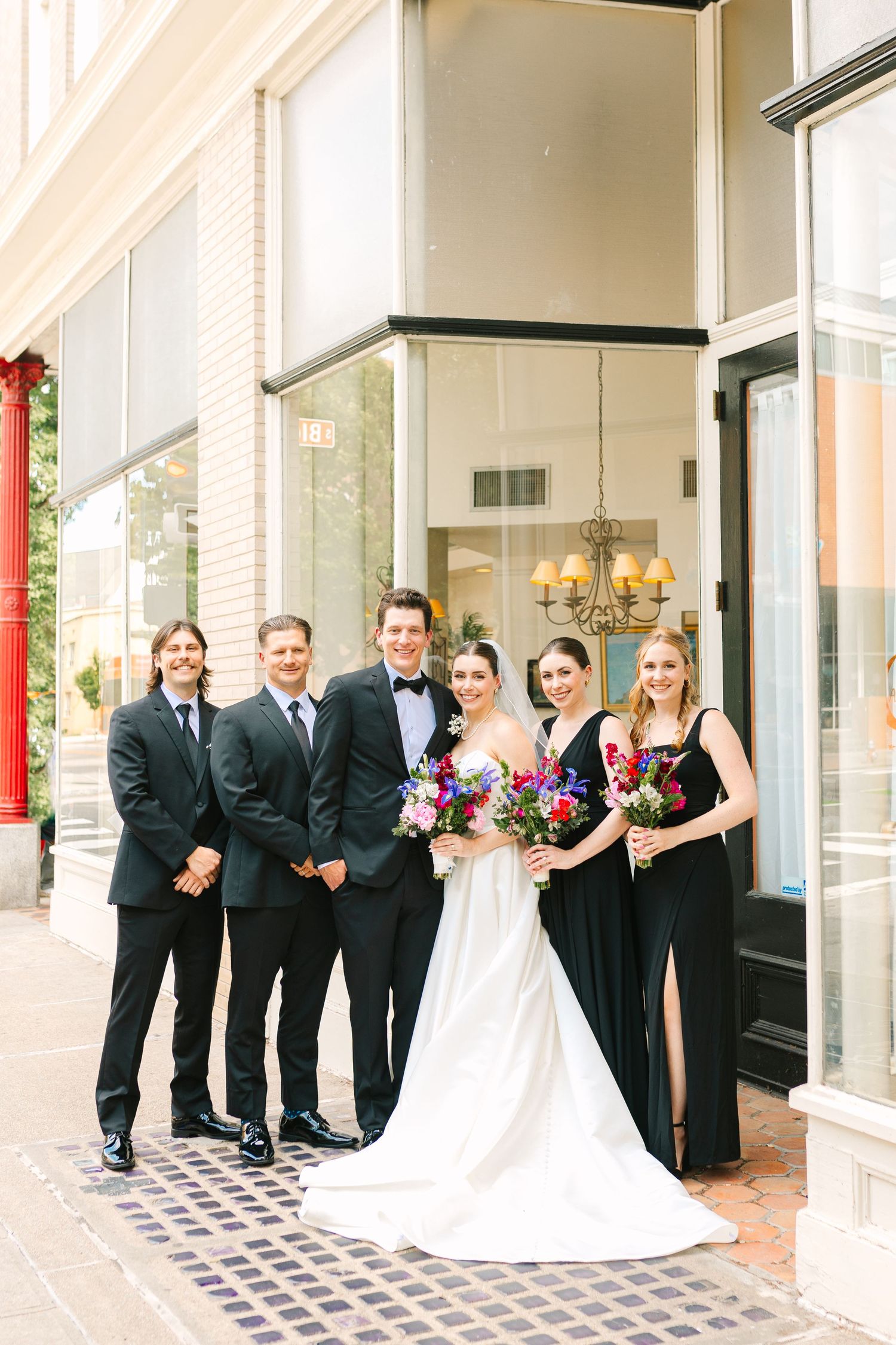 Wedding party in formal attire posed outside modern building with glass windows.