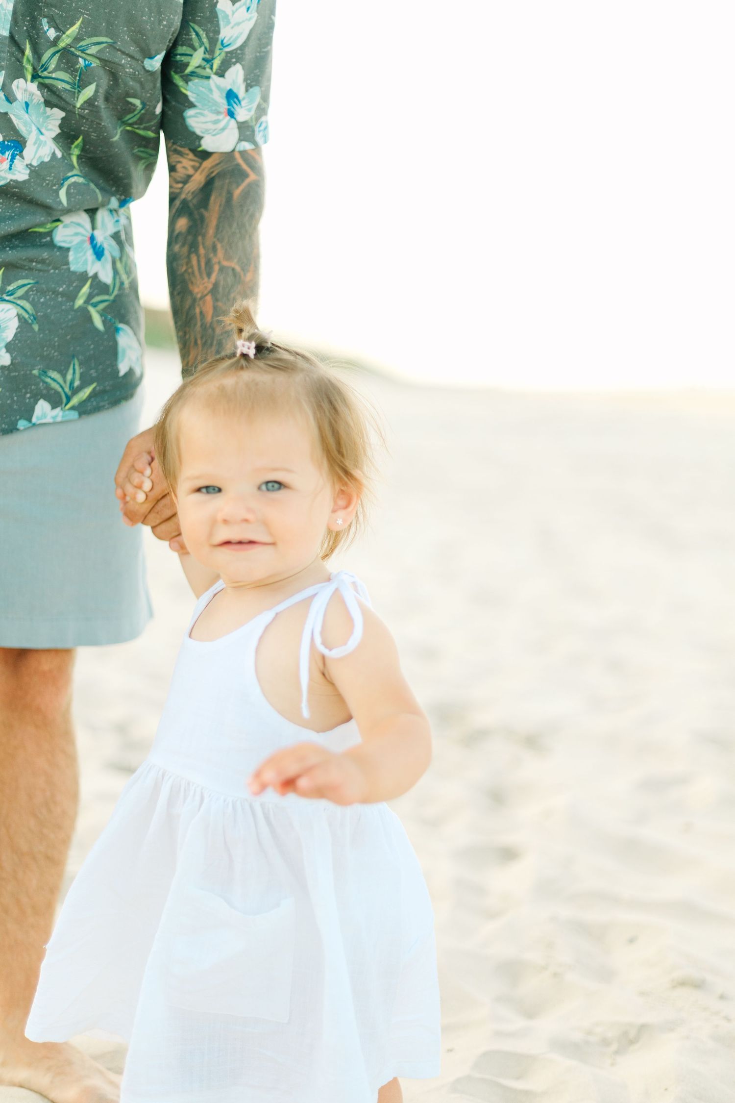 Young child in white dress playing on sandy beach with family.