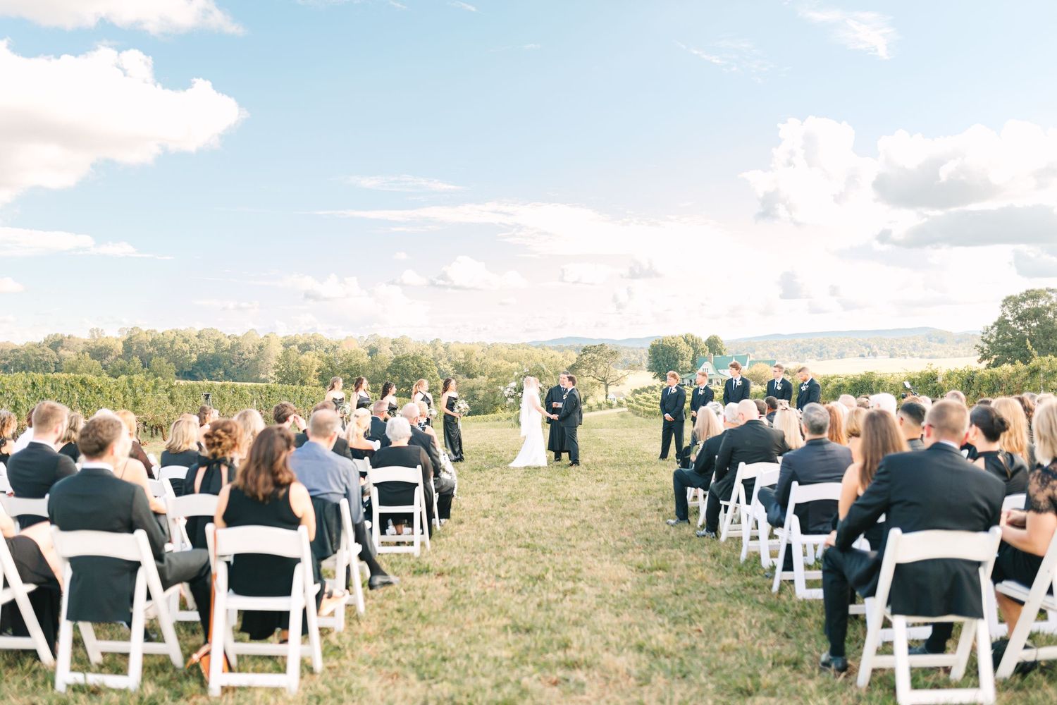 Outdoor wedding ceremony setup with white chairs in a scenic field under blue skies.