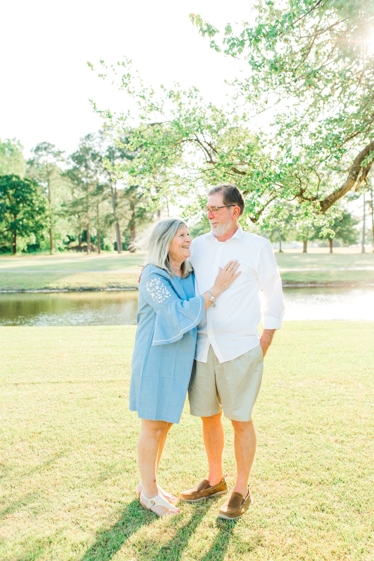 A couple shares a tender moment while standing together on a sunny lawn near a lake.