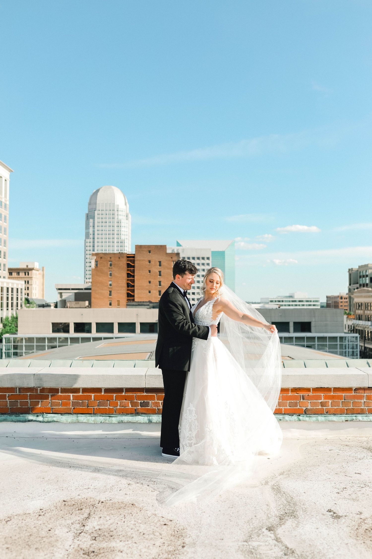 A couple in wedding attire poses on a rooftop with city buildings and blue sky in the background.