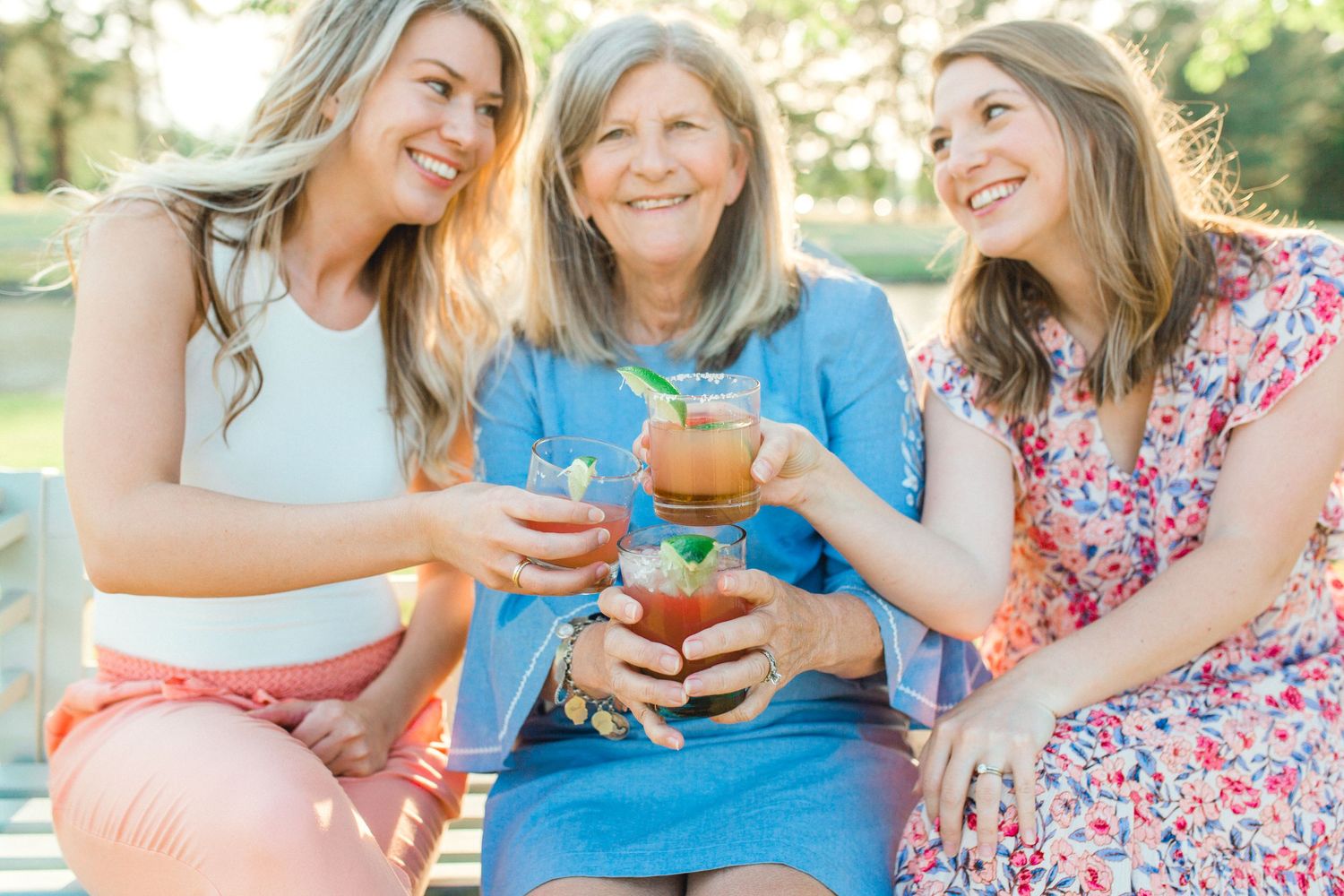 Three people share drinks and laughter while sitting outdoors in casual summer attire.