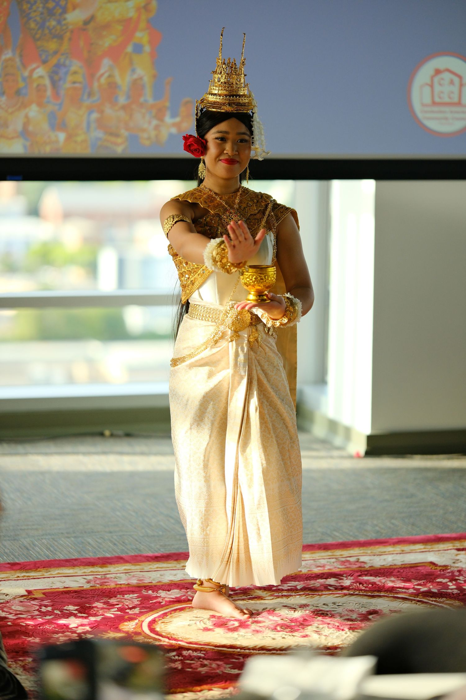 Traditional Thai dancer performing in elegant gold costume and ornate headdress on red carpet.