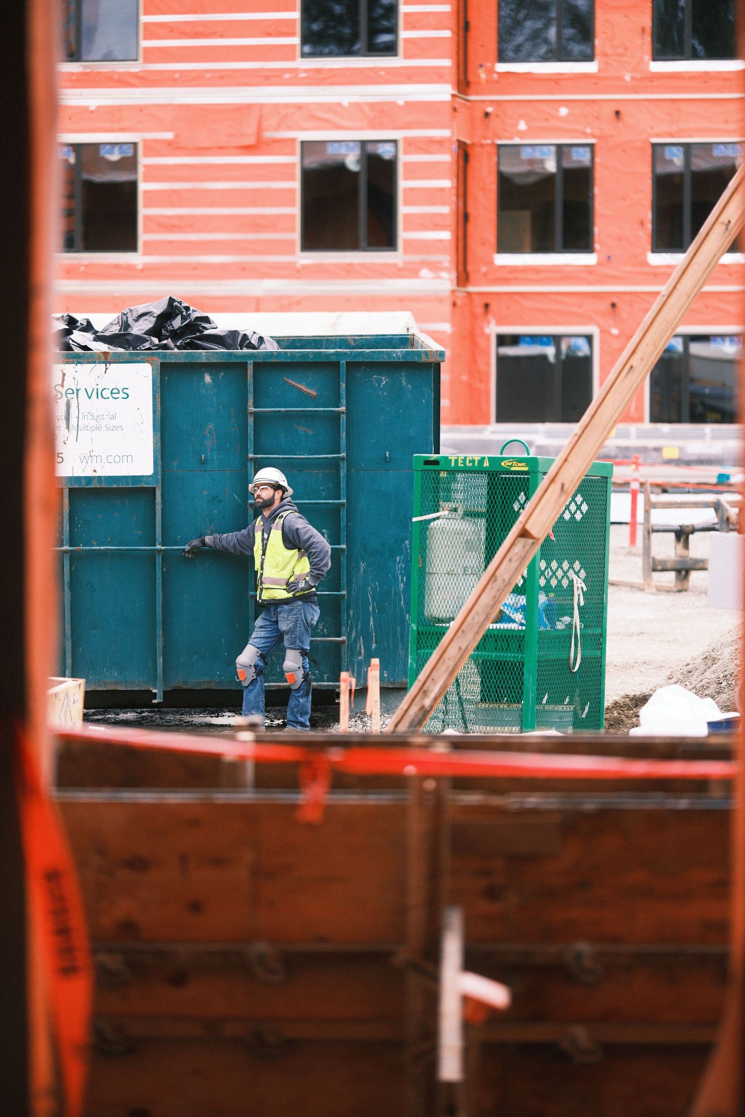 Industrial construction site with red walls and green dumpsters visible through window frames.