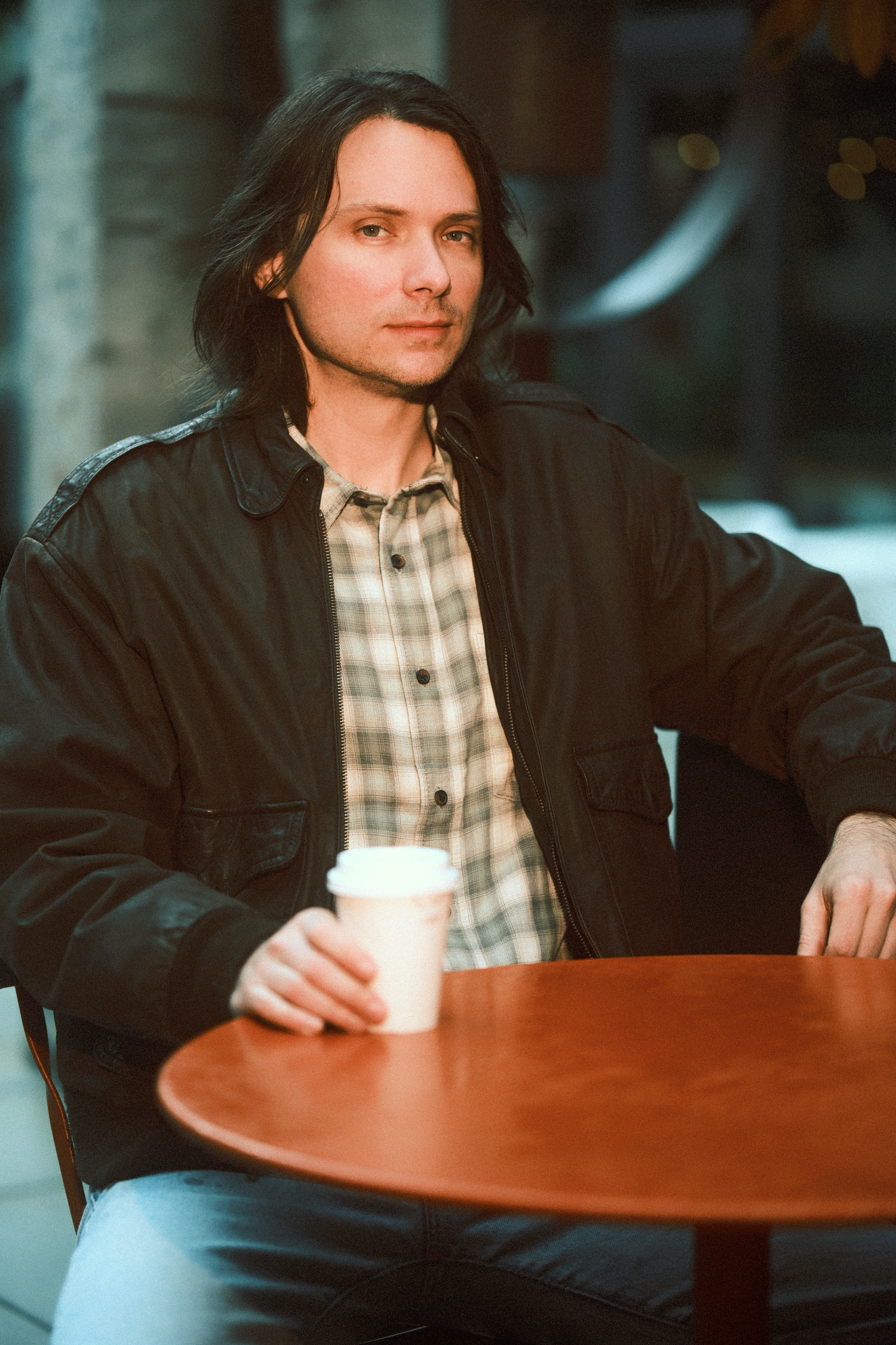 Person at cafe table with coffee cup wearing plaid shirt and dark jacket.