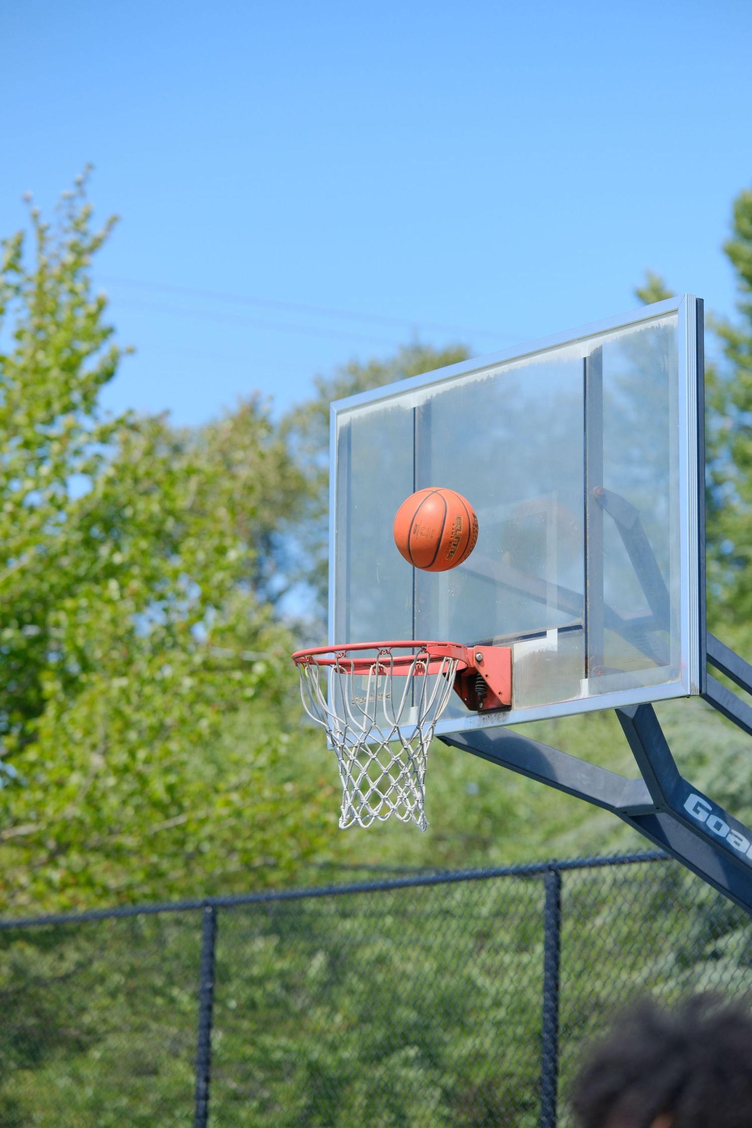 Basketball mid-air above hoop with clear backboard against blue sky.
