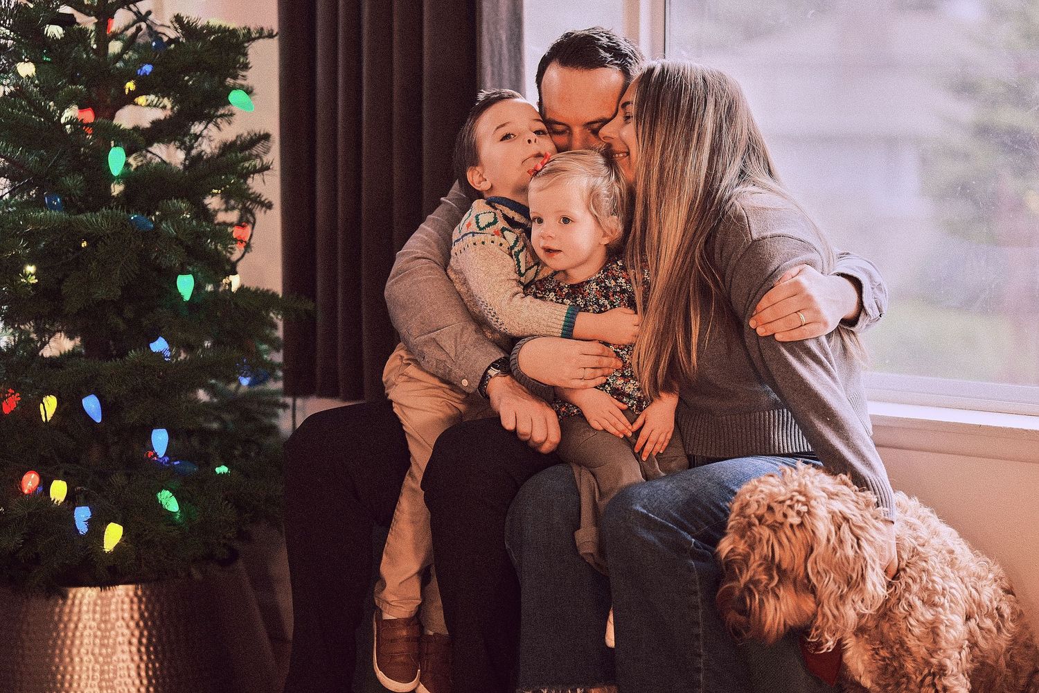A cozy family moment by a Christmas tree with a fluffy goldendoodle dog looking on.