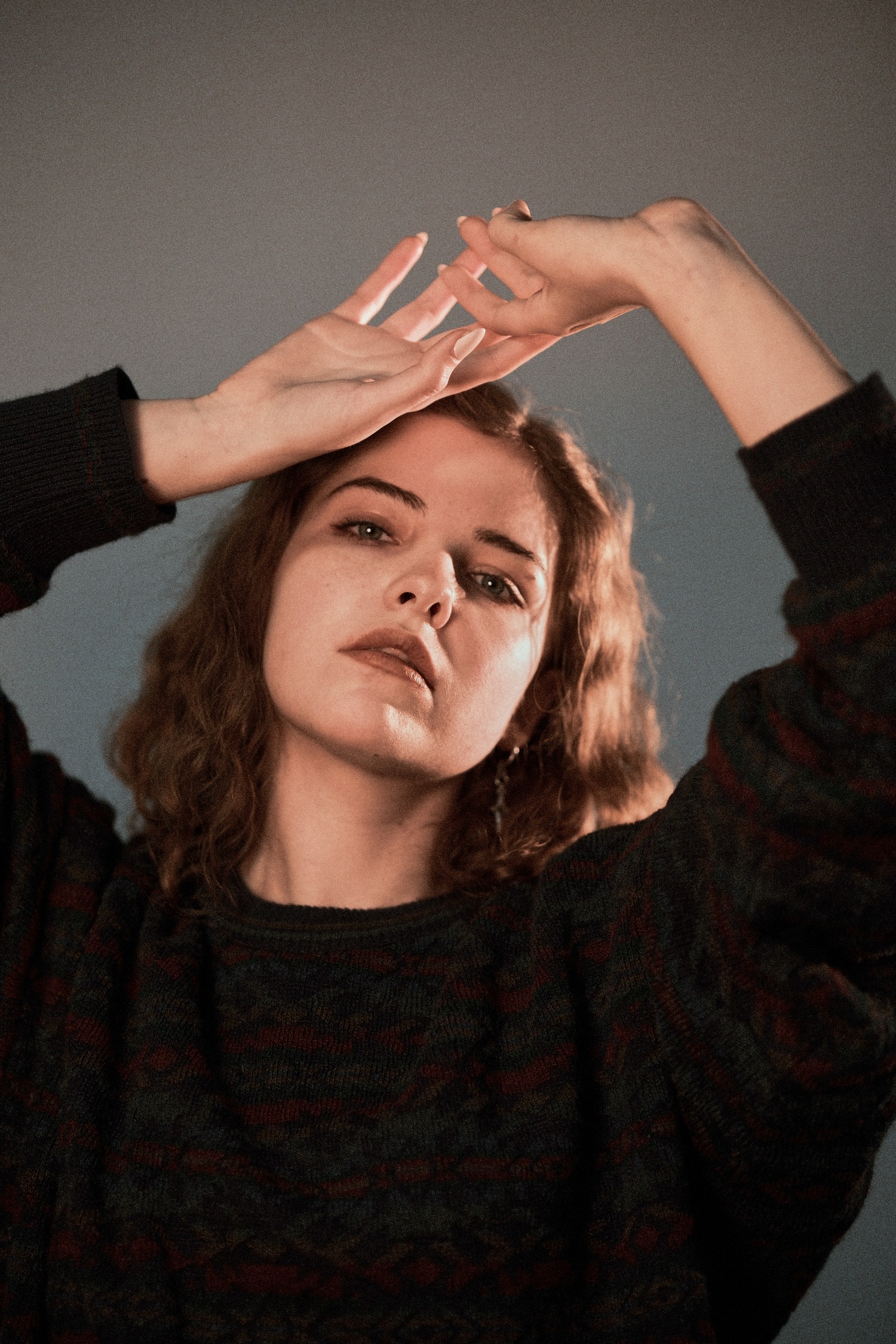Artistic portrait with hands raised in dramatic pose against neutral background.