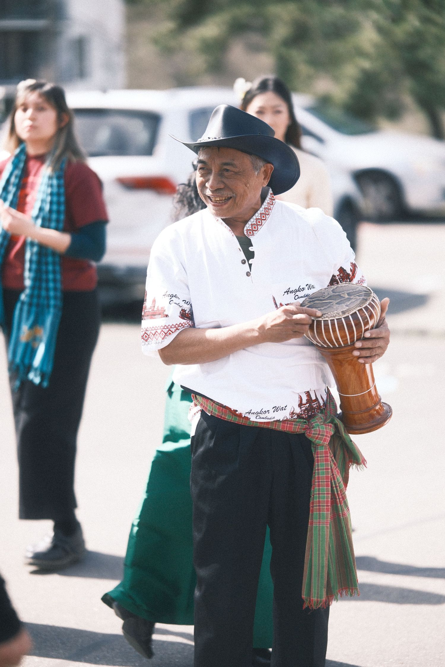 Musician in a cowboy hat and embroidered shirt holding a traditional drum instrument outdoors.