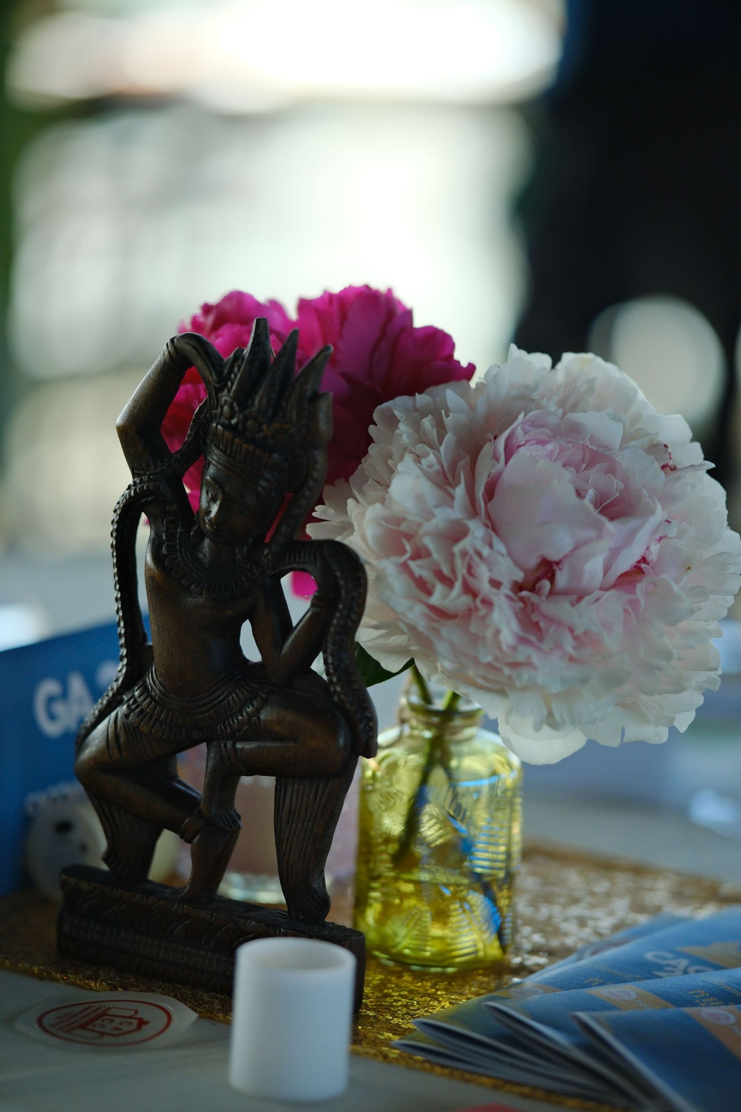 Decorative wooden statue next to pink peony flower in yellow glass vase.