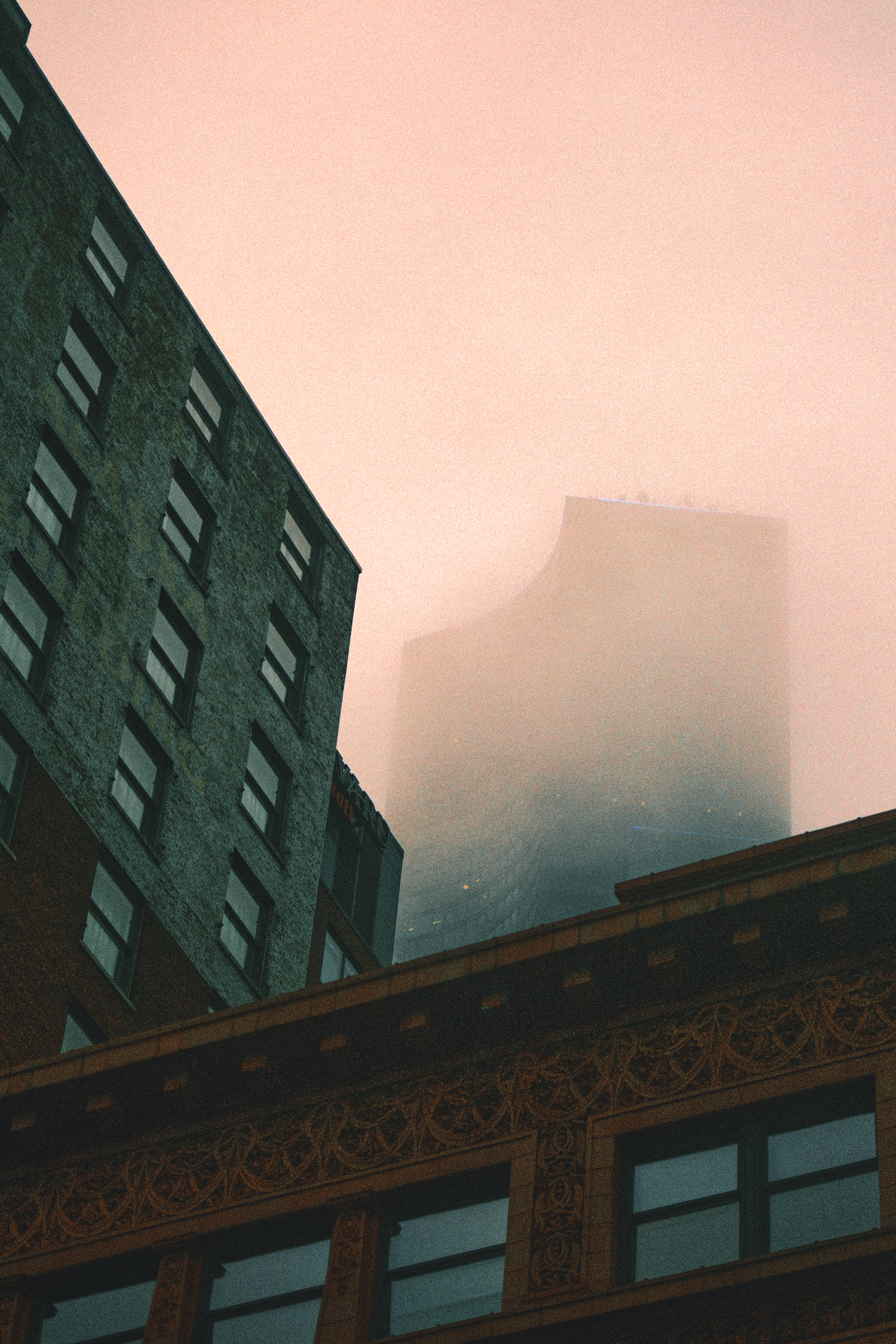 Urban buildings with ornate facade against pink sky with fog obscuring upper floors.