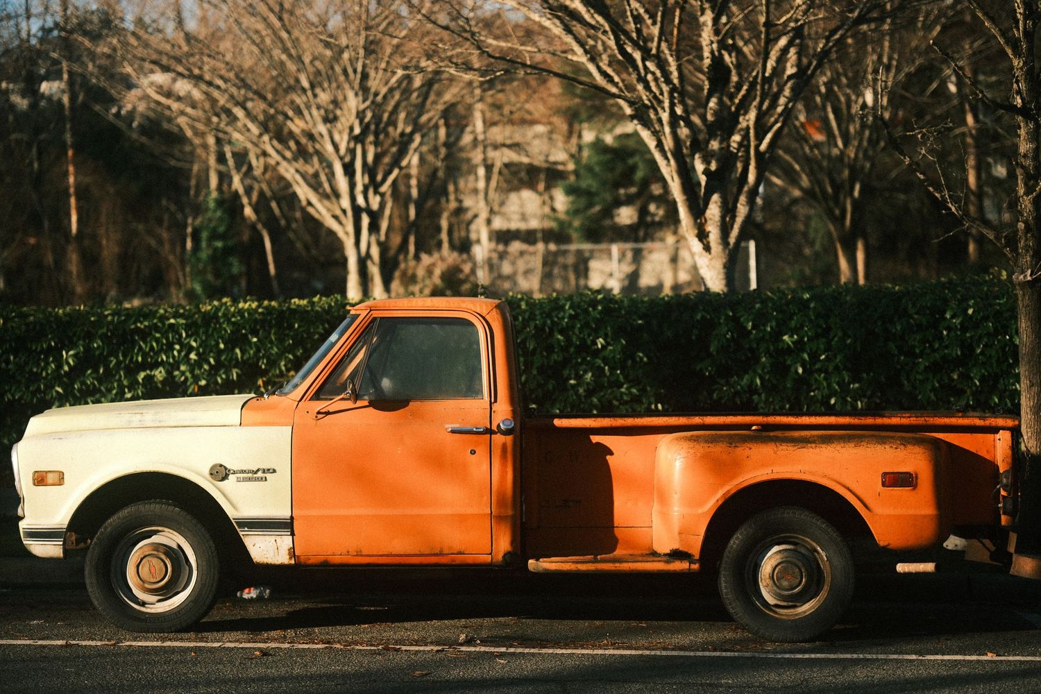 Vintage orange pickup truck parked alongside other vehicles on a tree-lined street.