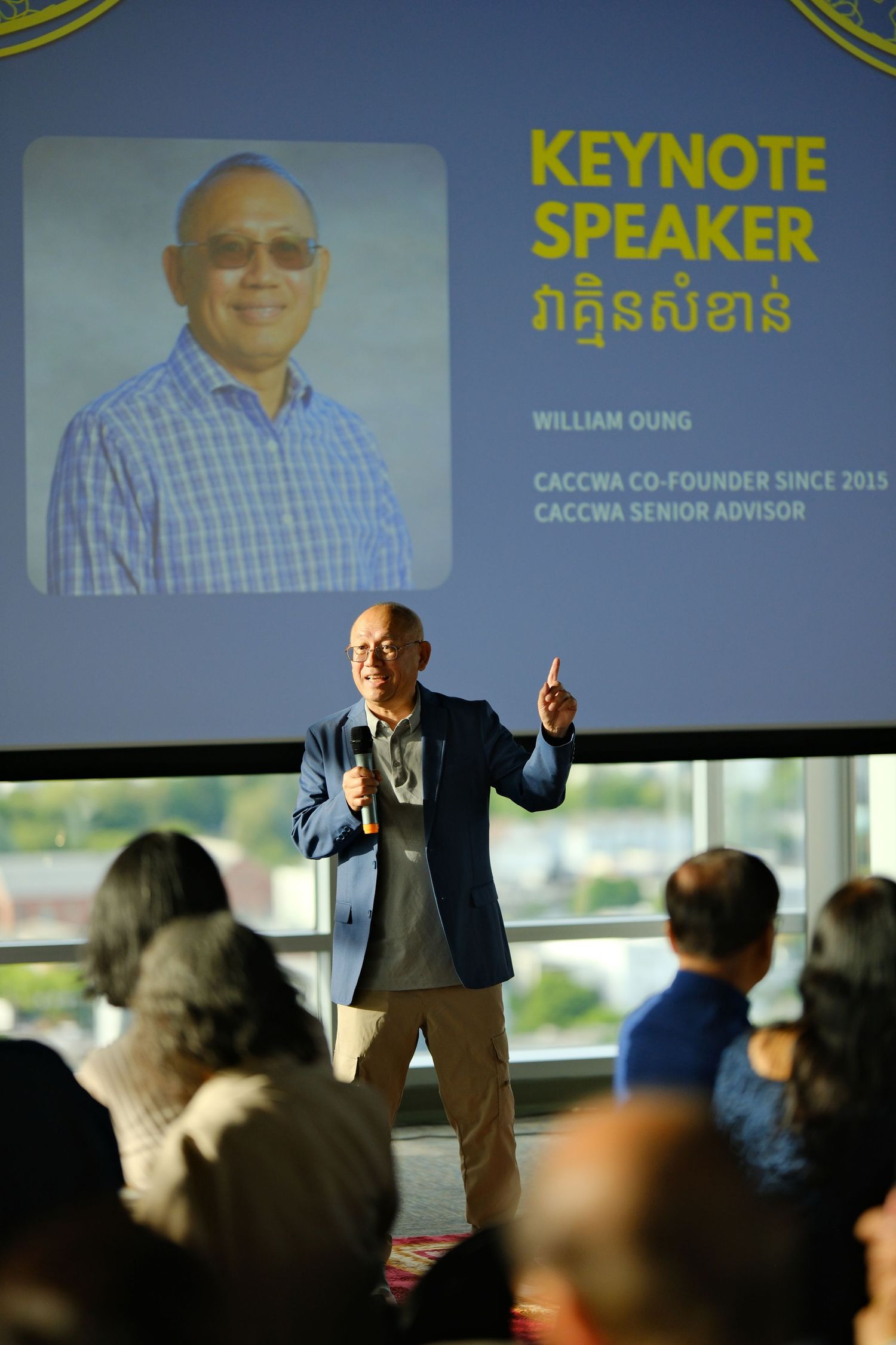 A keynote speaker presents to an audience at a business conference with yellow text displayed on screens behind them.