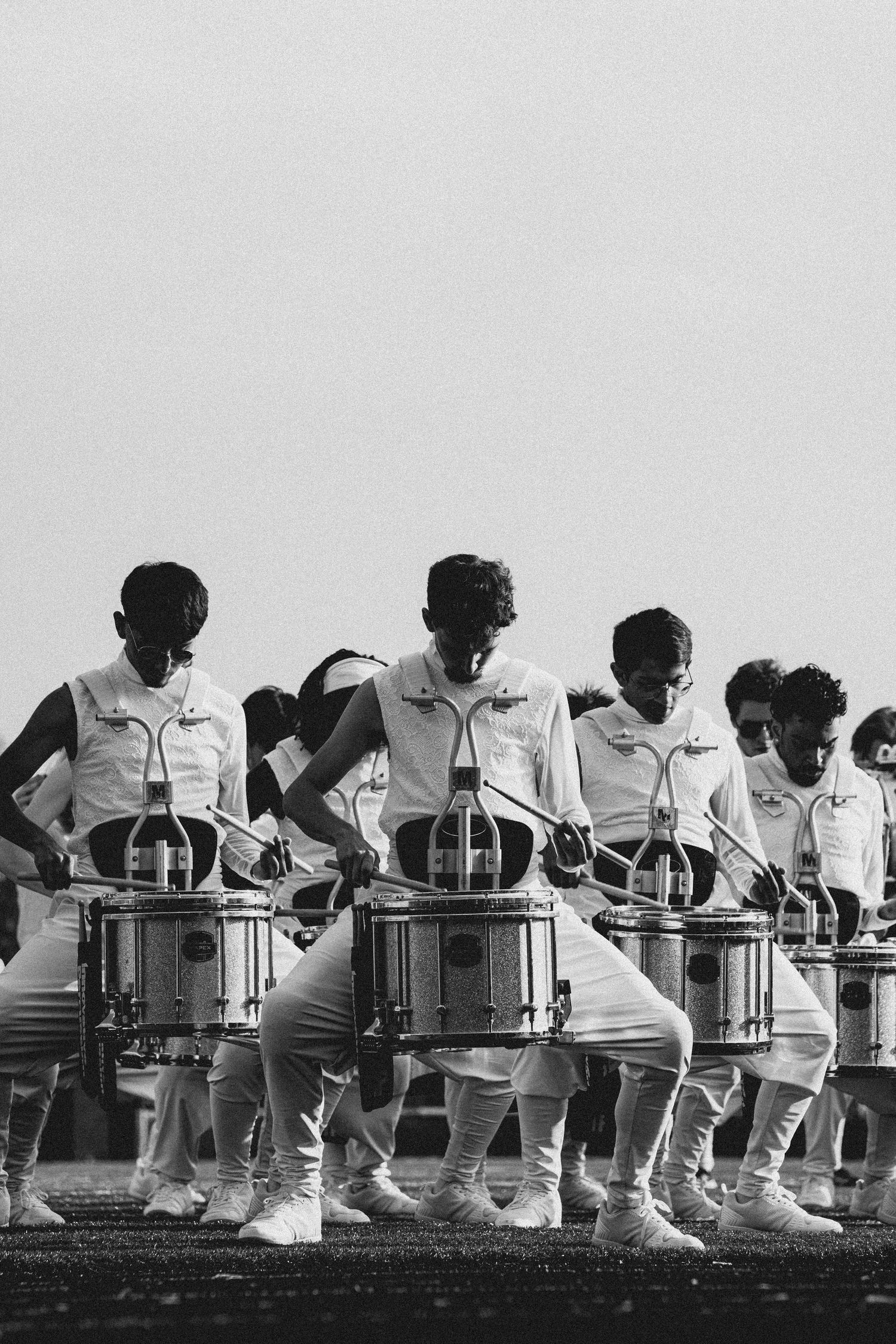 Black and white photo of a marching band drumline performing in white uniforms, playing snare drums in synchronized formation.